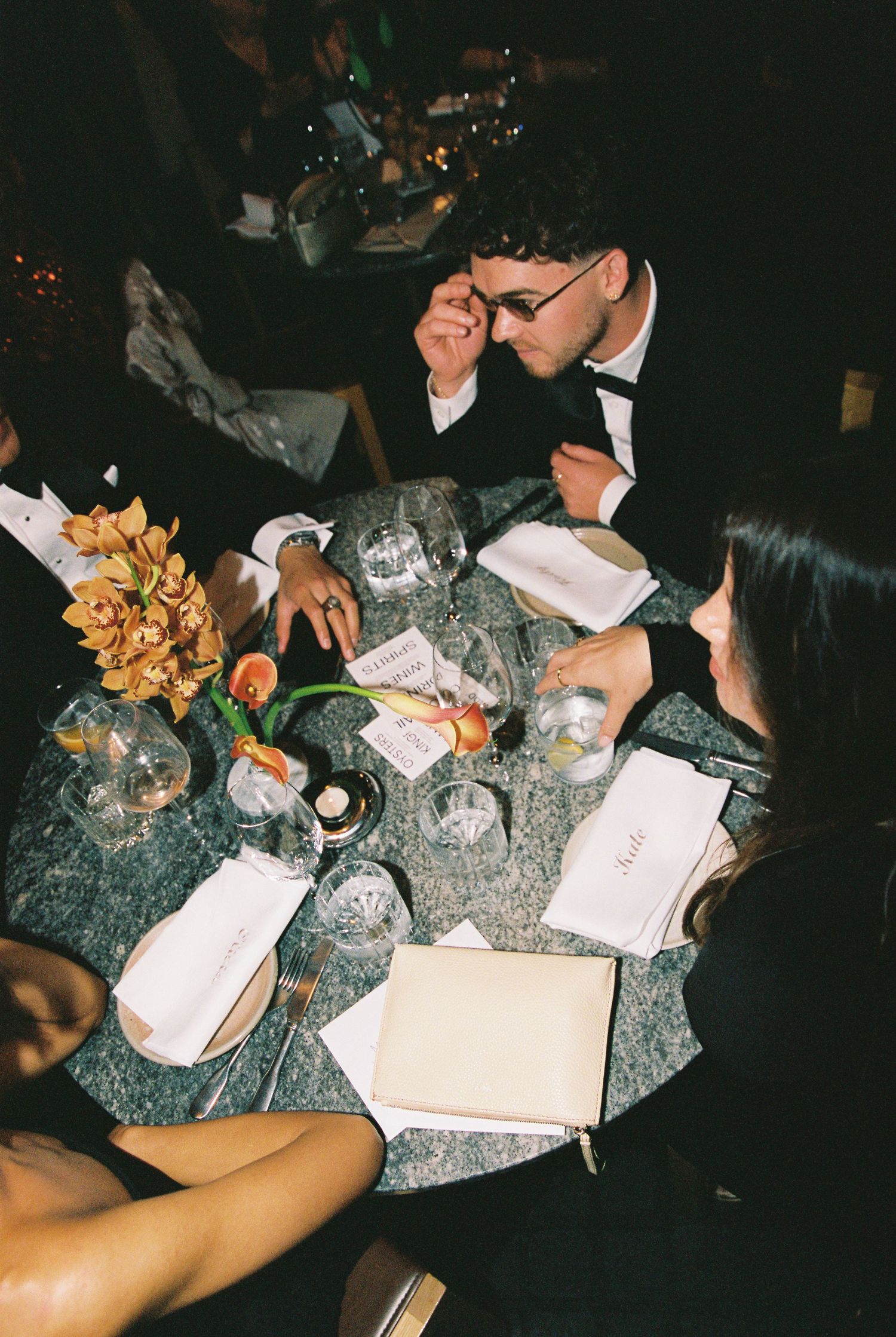Guests in formal attire seated at an elegant wedding reception table with floral centerpieces and place settings.