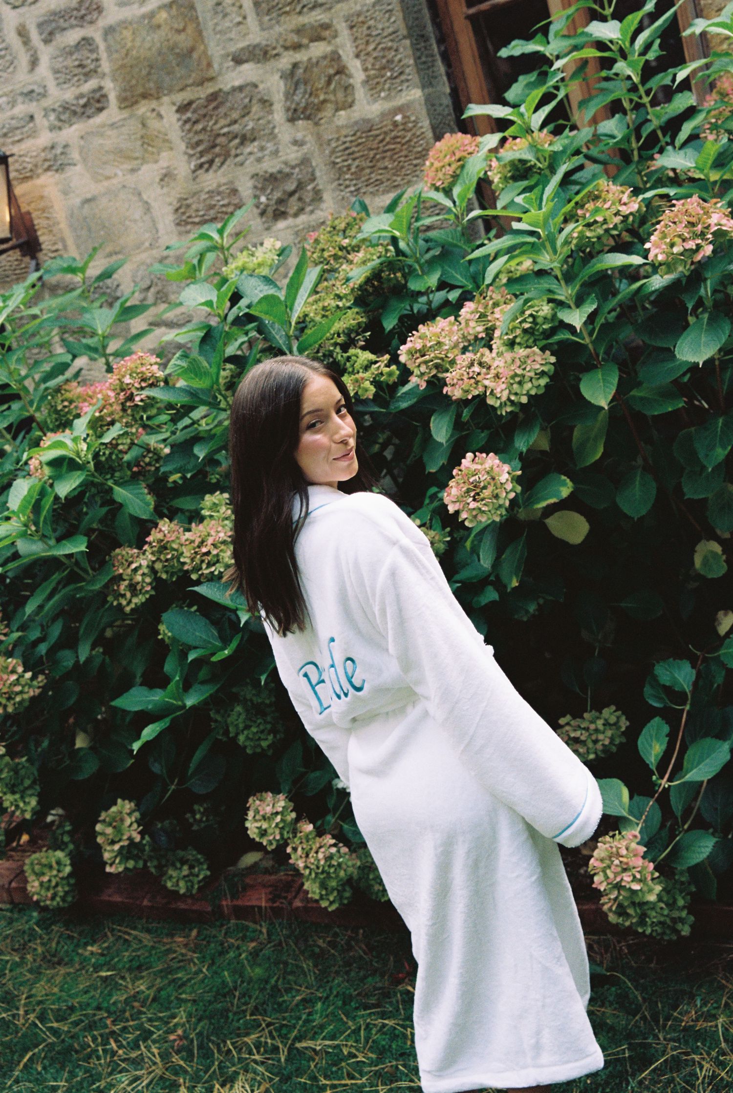 Bride in white monogrammed robe poses in front of blooming hydrangea bushes at stone venue.