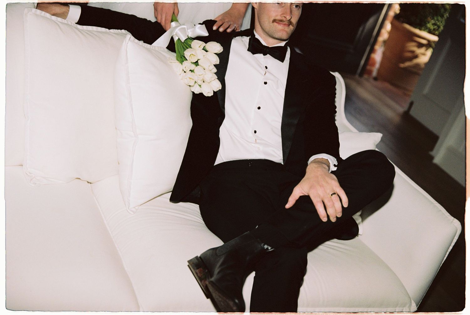 Groom in black tuxedo relaxing on white sofa holding bridal bouquet of white roses.