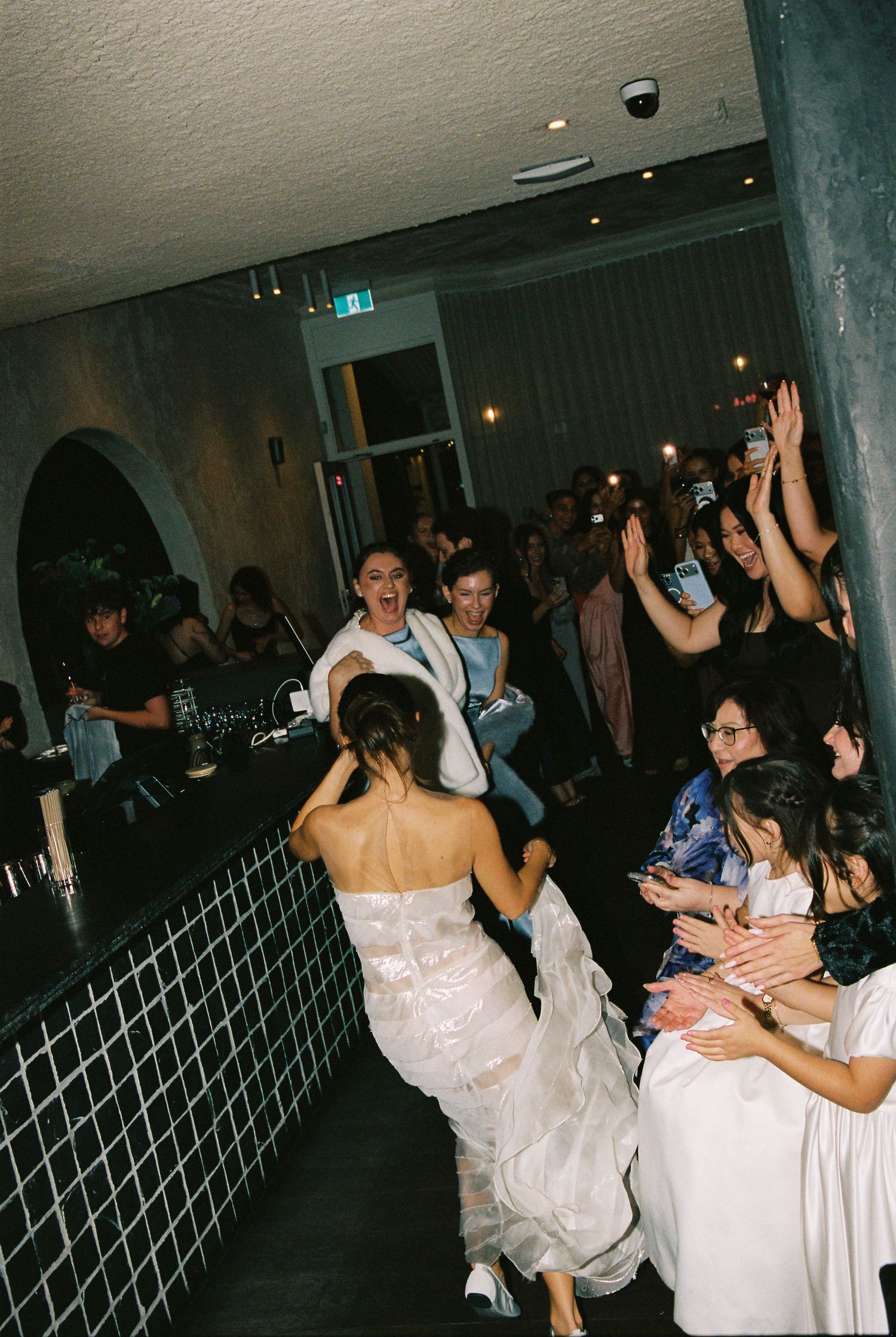 Bride in white wedding dress energetically dancing with cheering guests at reception party.