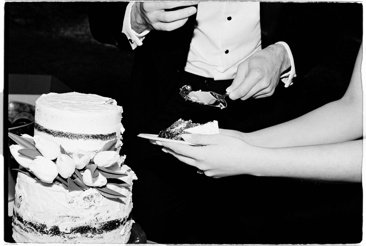 Black and white photo of a bride and groom cutting a tiered wedding cake decorated with flowers.
