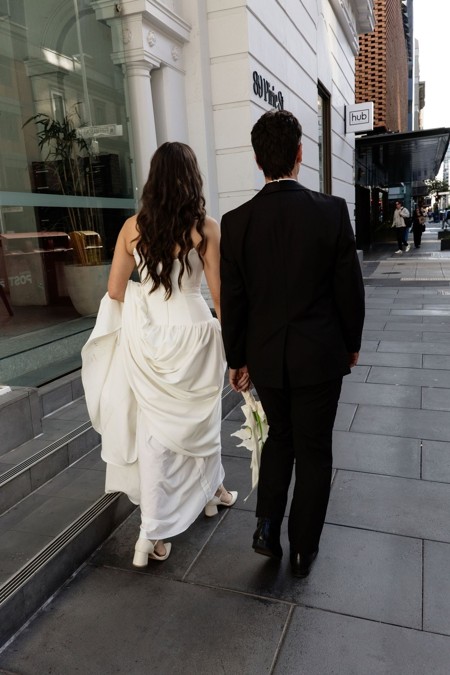 Bride in white gown and groom in black tuxedo walking hand in hand down a city street.
