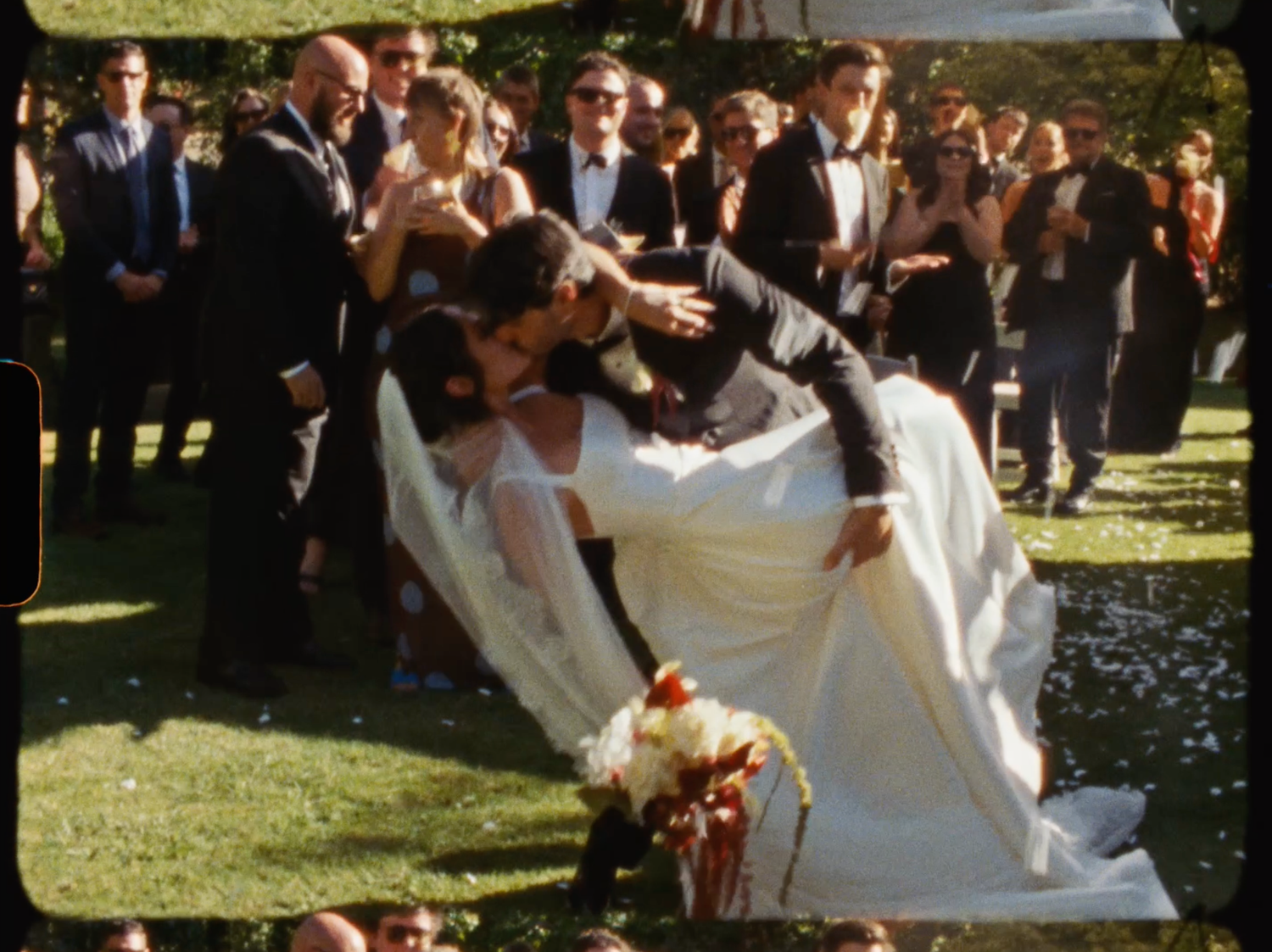 Bride and groom share a romantic dip kiss surrounded by cheering wedding guests on a green lawn.