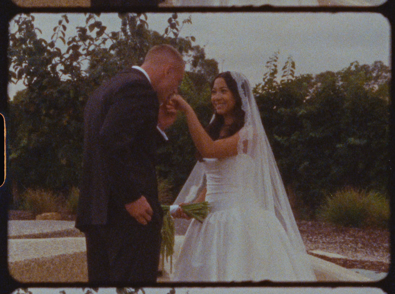 Bride smiling as groom kisses her hand in romantic outdoor garden wedding portrait.