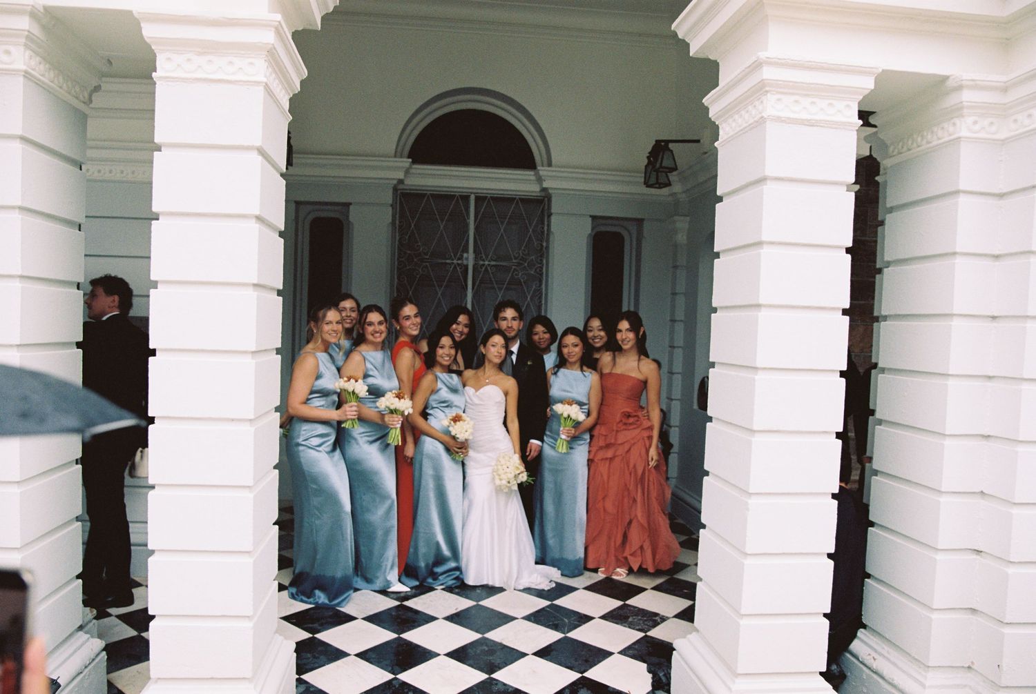 Bride in white gown poses with bridesmaids in blue and pink dresses on checkered floor under arches.
