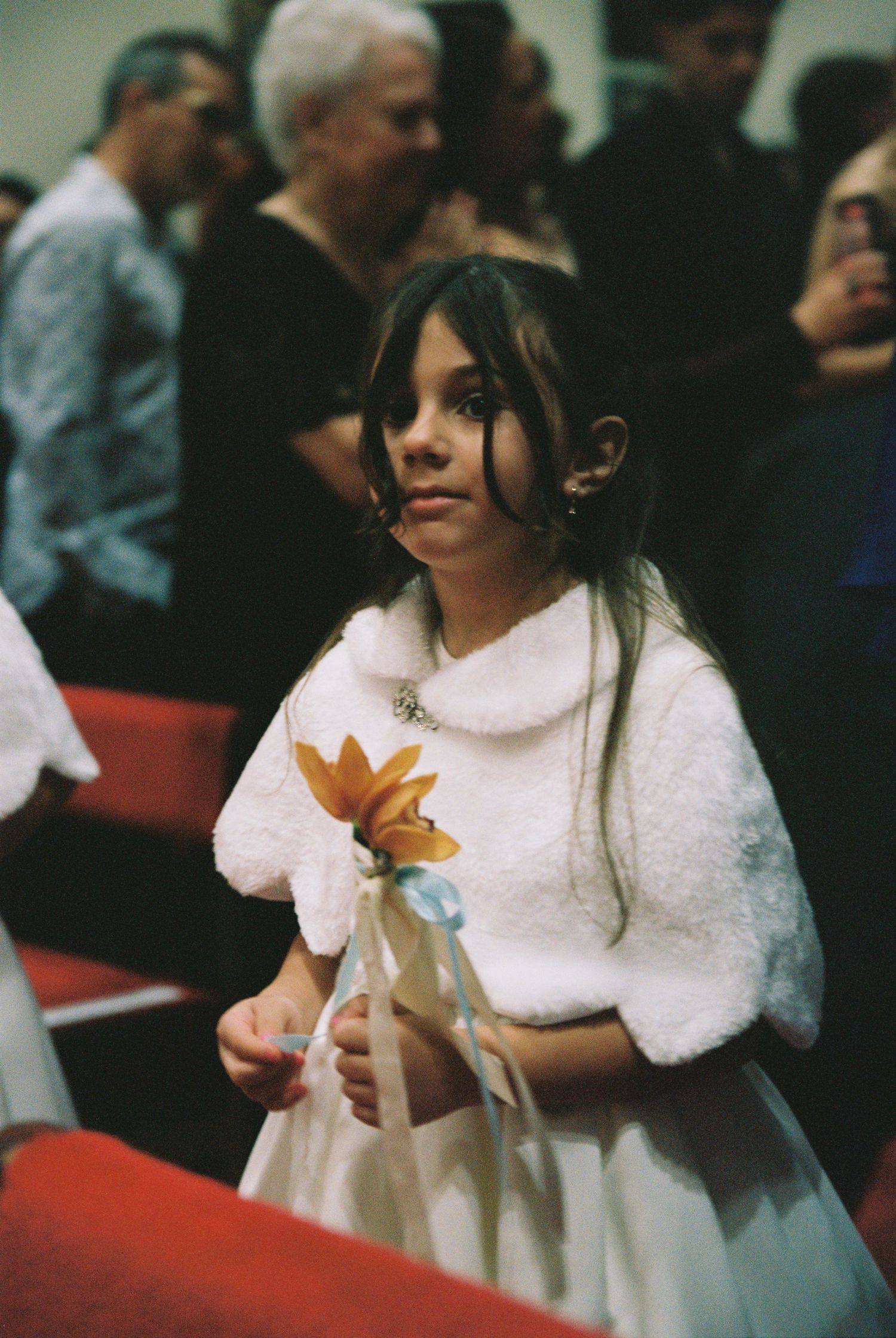 Young flower girl in white dress and fur shrug holds yellow flower during wedding ceremony indoors.