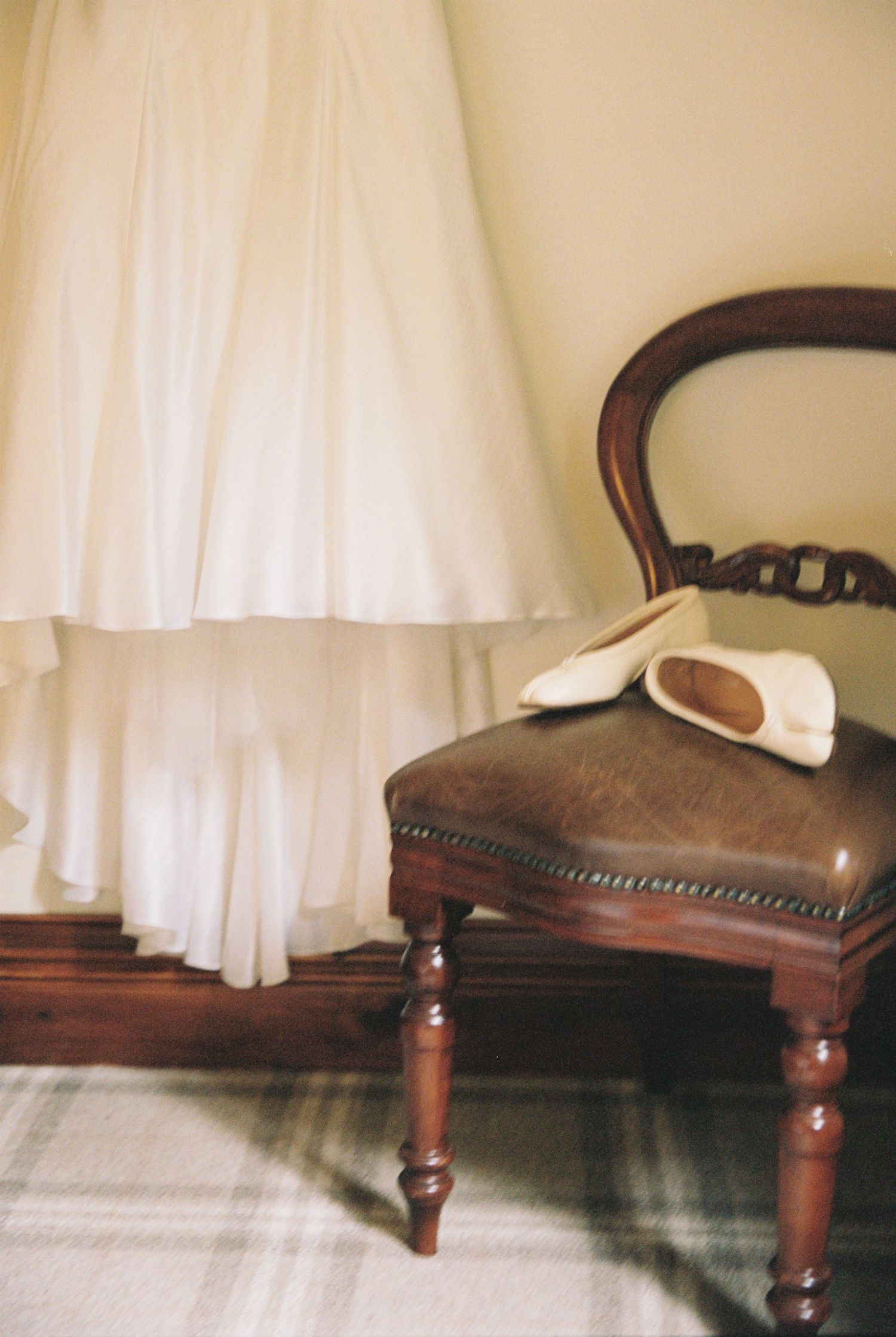 Elegant white wedding dress hem beside vintage wooden chair holding bridal shoes on tartan floor.