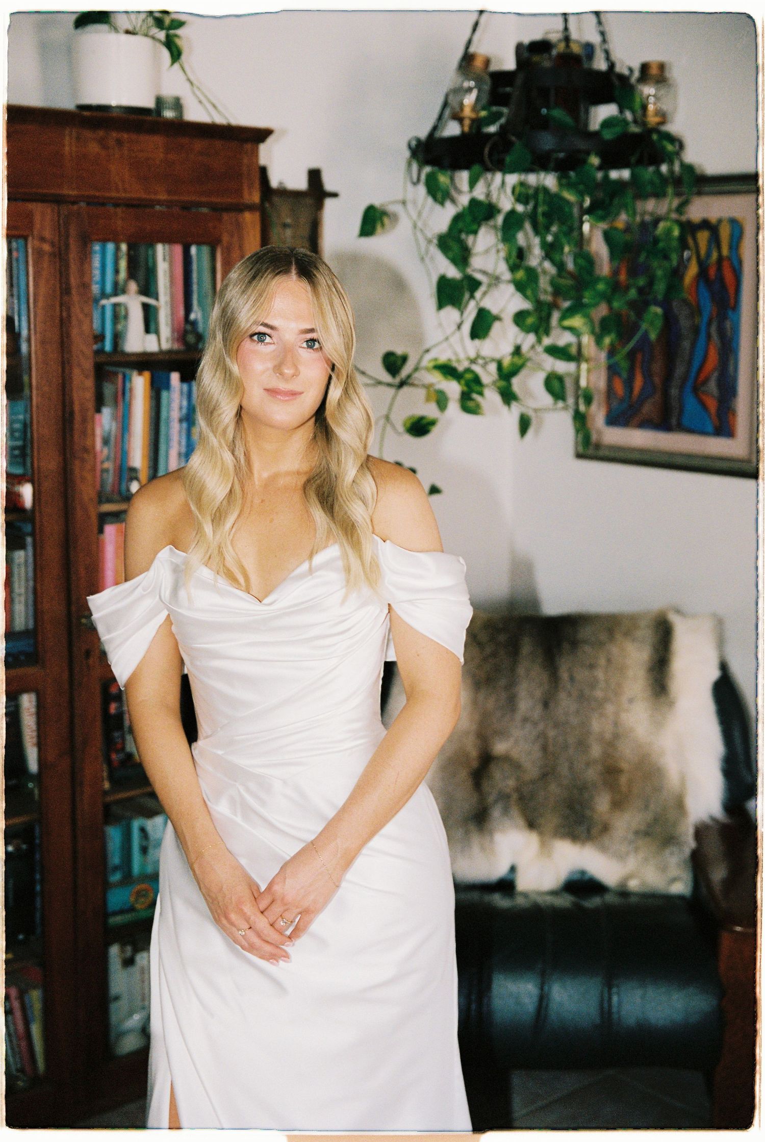 Elegant bride in white off-shoulder fitted gown standing in room with bookcase and plants.
