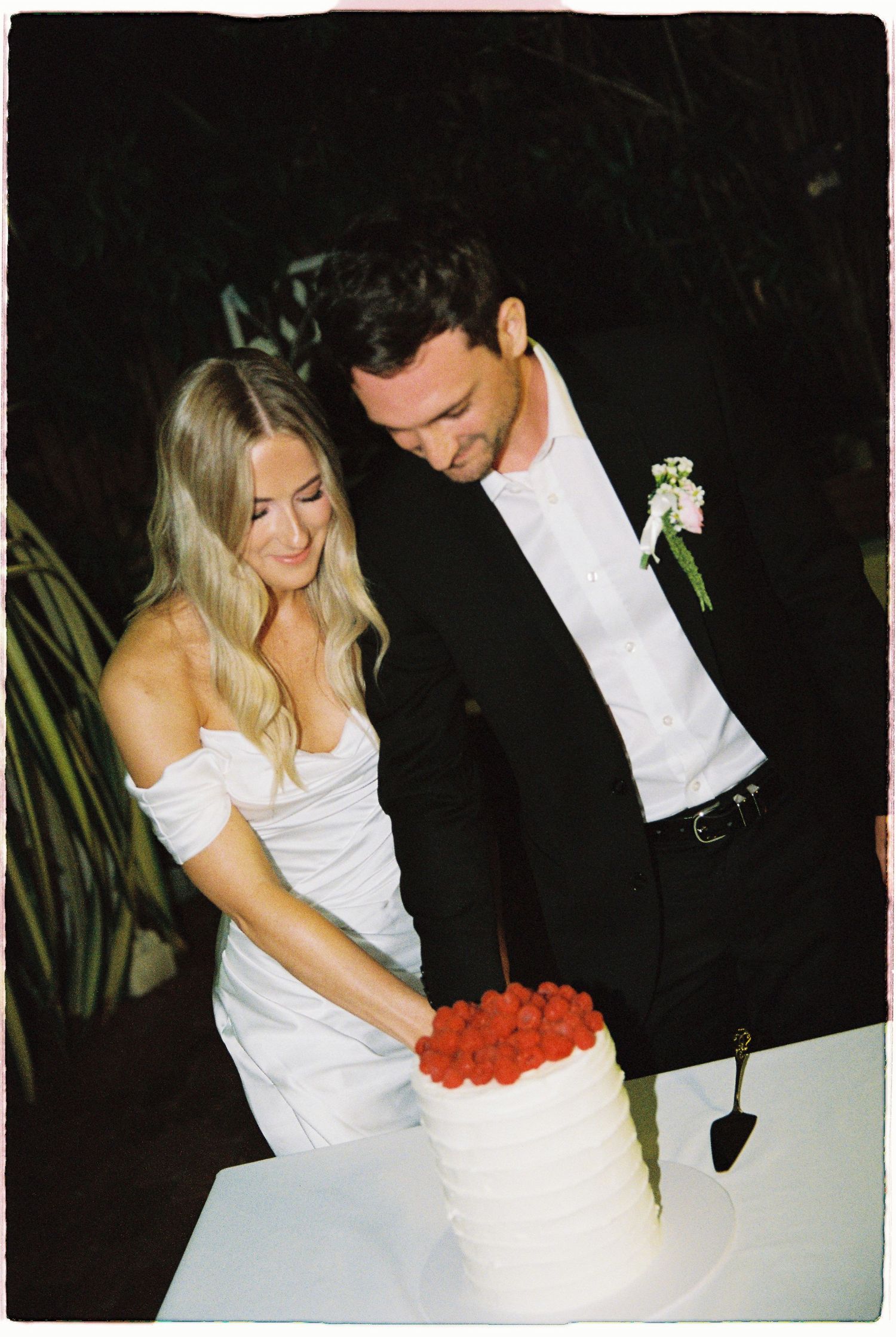 Smiling bride in white off-shoulder dress and groom in black suit cutting raspberry-topped wedding cake.