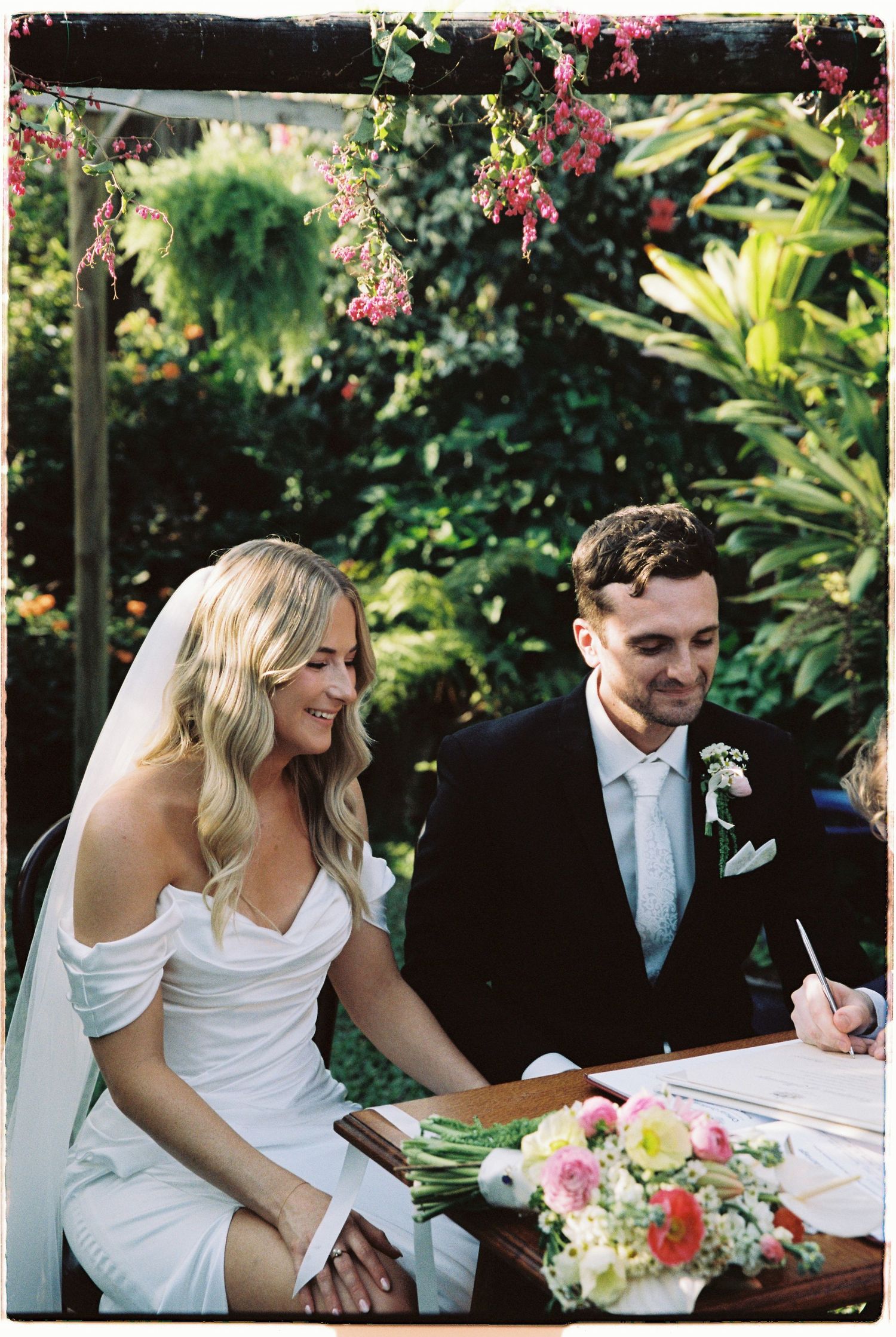 Smiling bride in off-shoulder gown signs marriage certificate beside groom in garden ceremony.