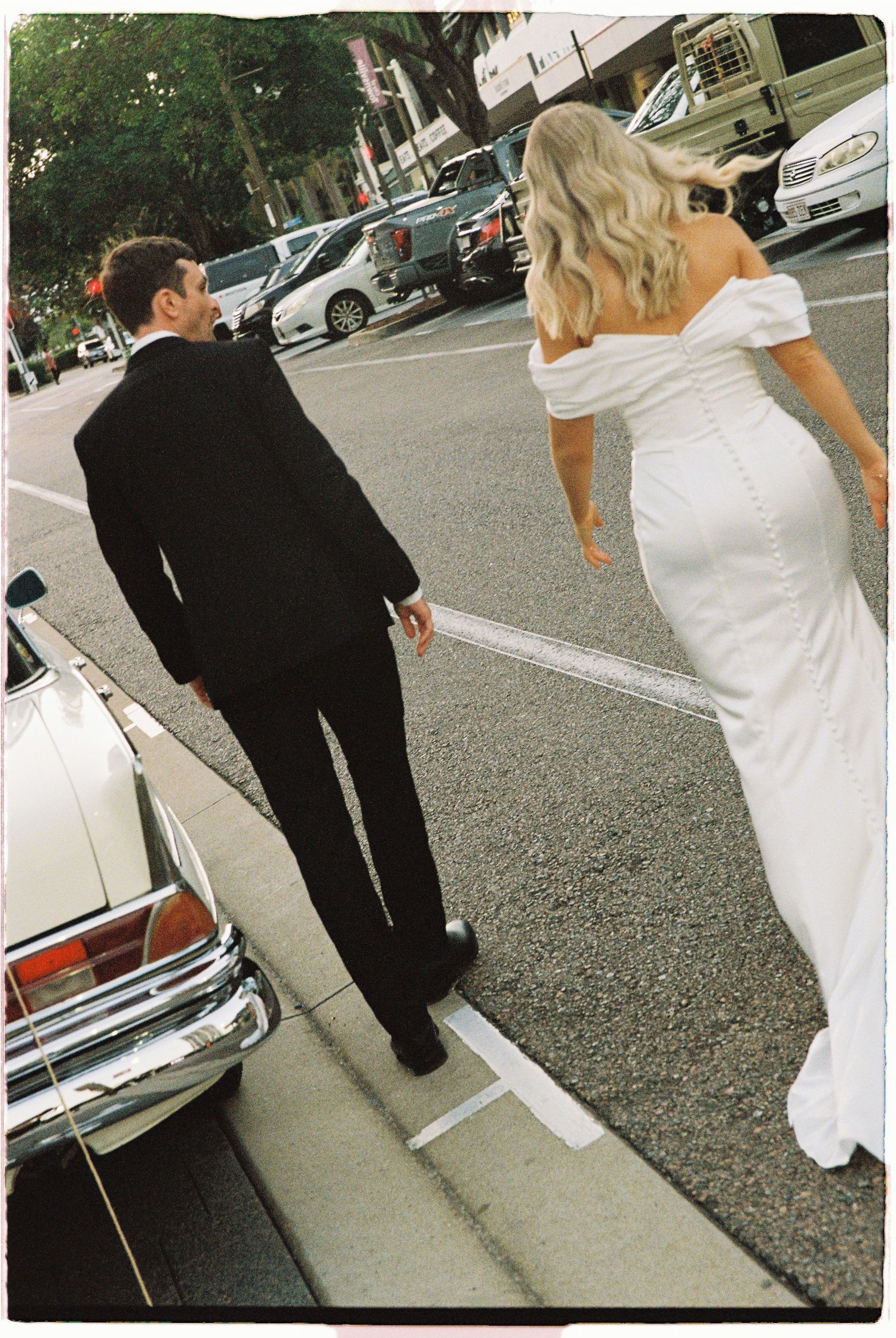 Bride in fitted off-shoulder gown and groom in black suit walk hand in hand near vintage car.