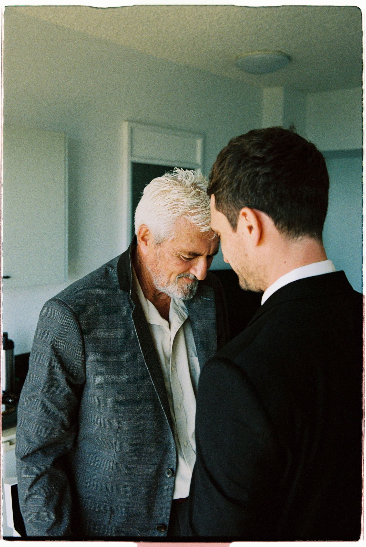 Older silver-haired man in gray suit shares an intimate moment with a younger man in a black suit indoors.