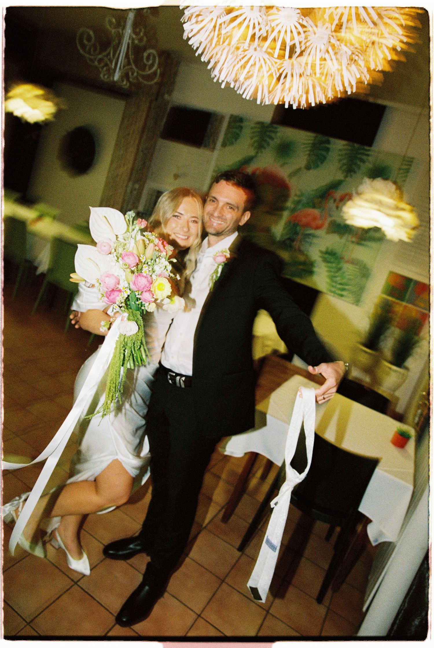 Smiling bride and groom pose together at wedding, bride holding colorful bouquet in restaurant venue.