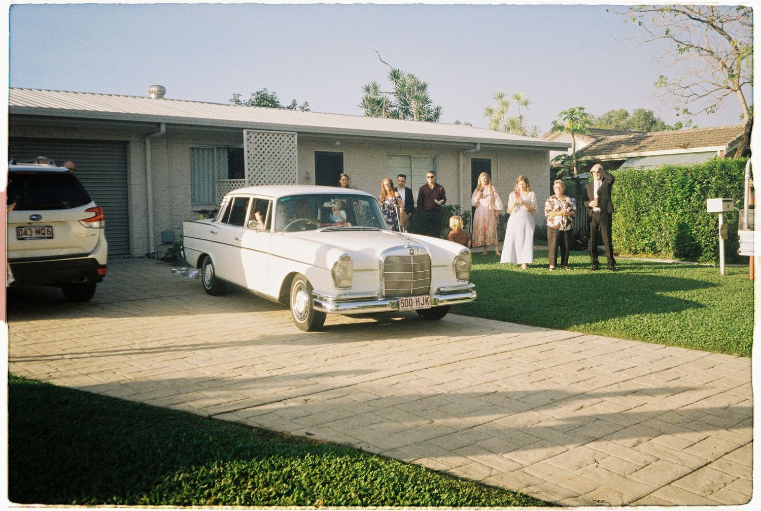 Vintage white Mercedes-Benz wedding car parked in driveway with wedding guests gathered outside a modern ranch-style home.