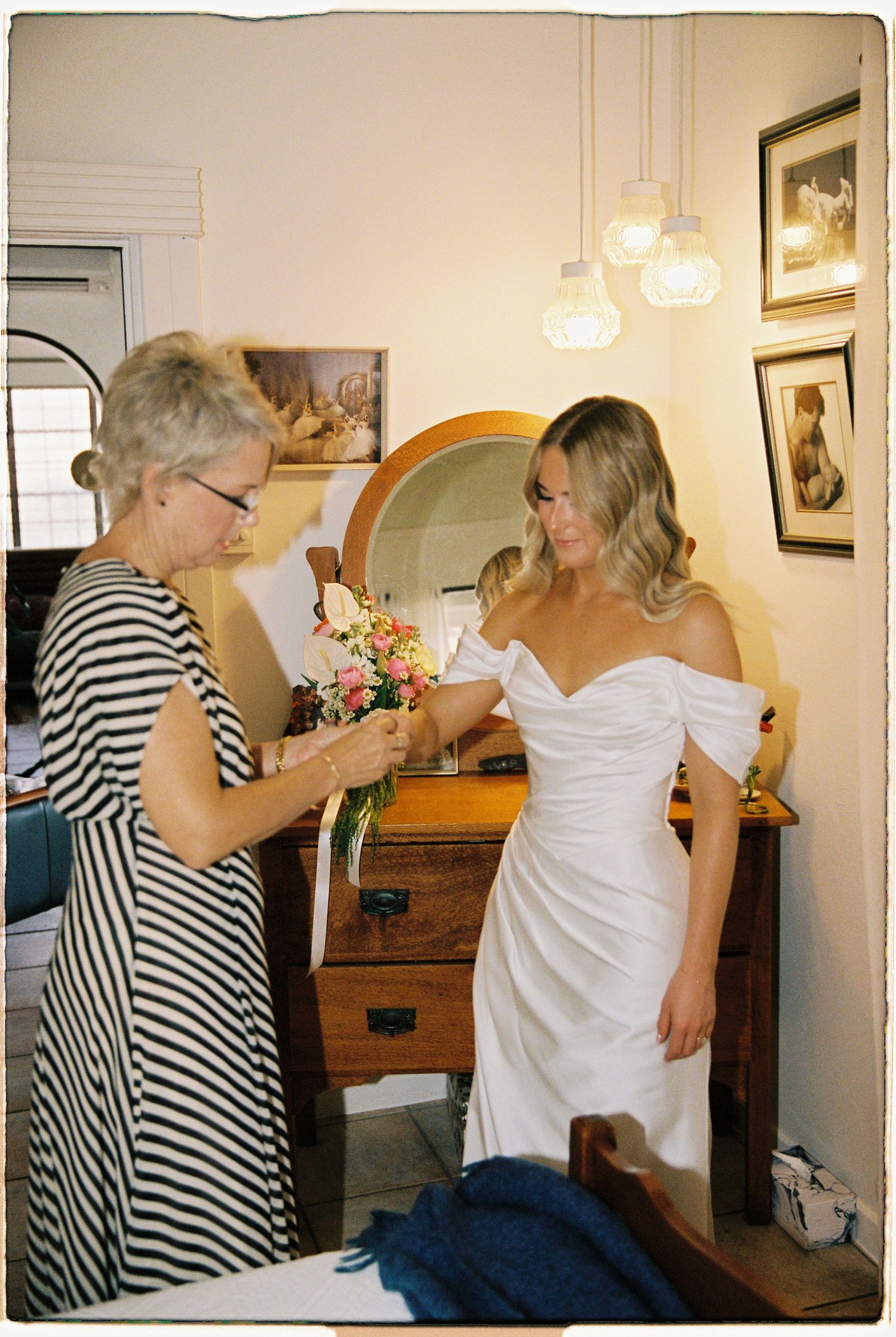 Bride in white off-shoulder gown receives bouquet of pink flowers from older woman in striped dress near vintage mirror.