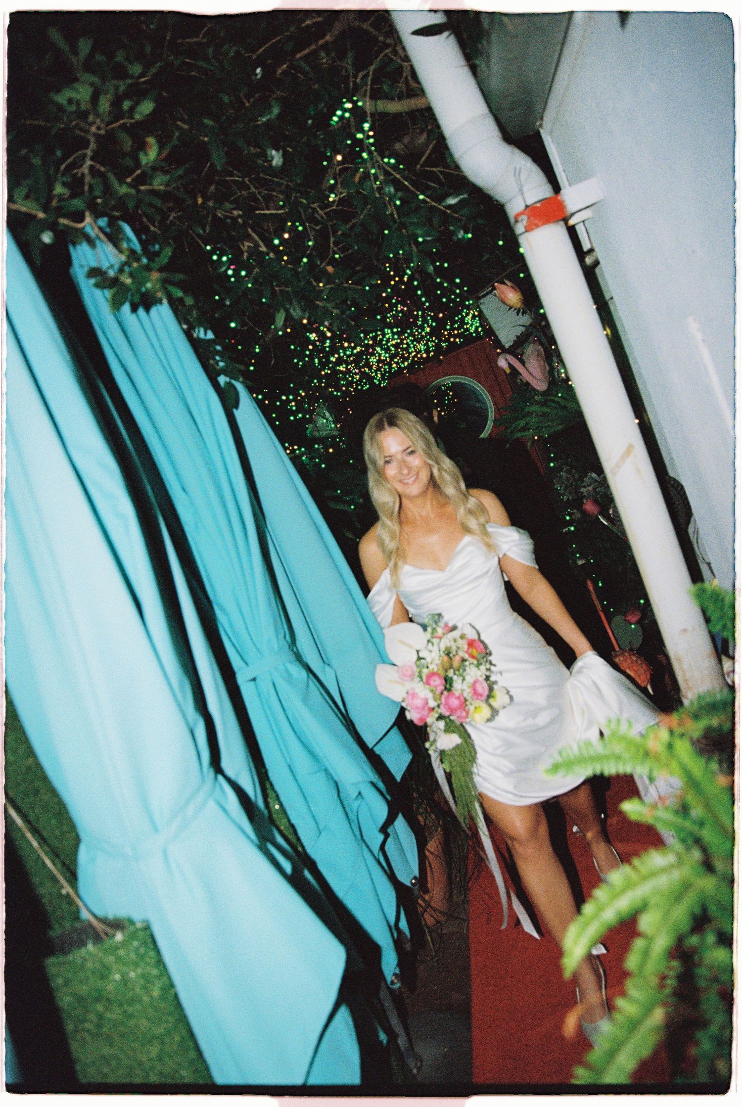 Bride in white off-shoulder mini dress holding pink bouquet walking through teal curtains at outdoor wedding.
