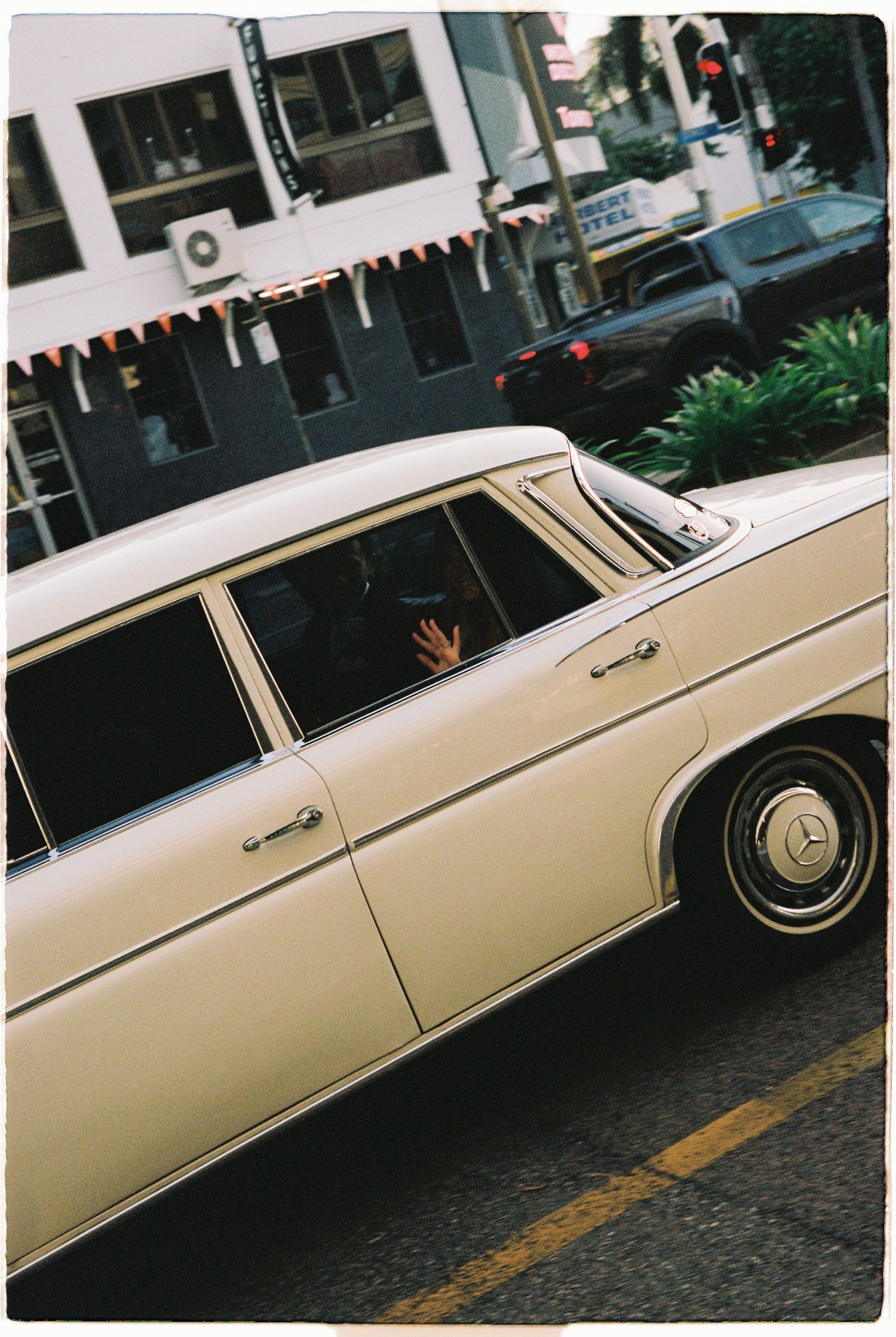 Classic cream Mercedes-Benz sedan with star emblem drives through urban intersection, passenger waving inside.