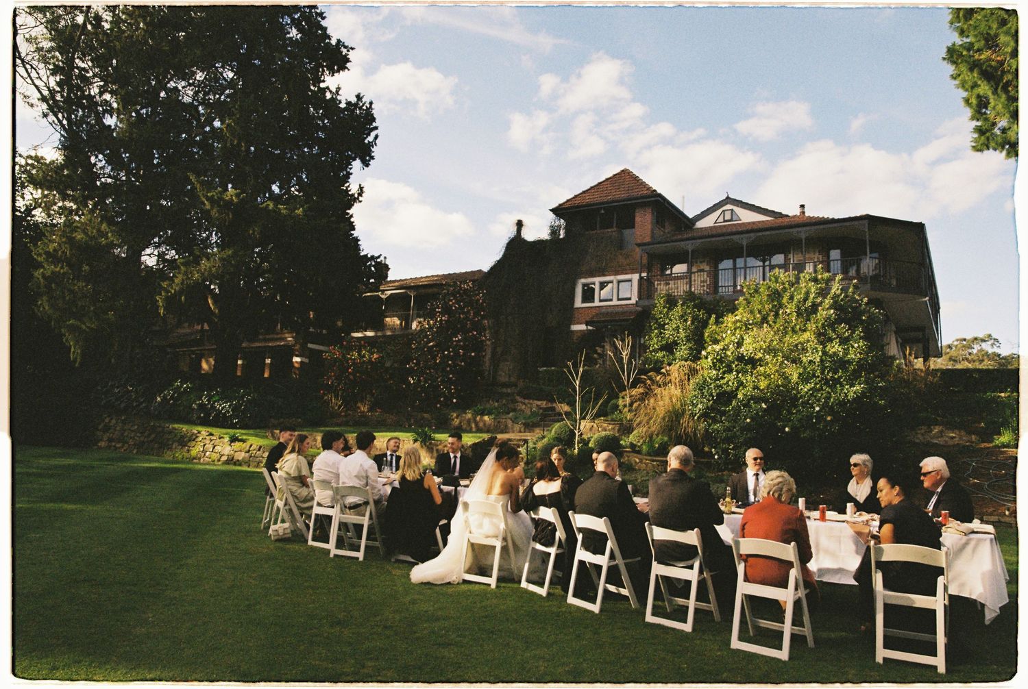 Wedding guests seated in white chairs on lawn during outdoor ceremony beside historic manor house.