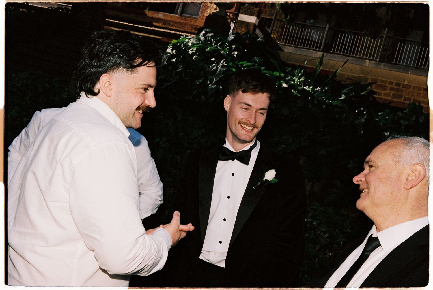 Three men in formal attire laughing and conversing at an outdoor wedding reception at night.