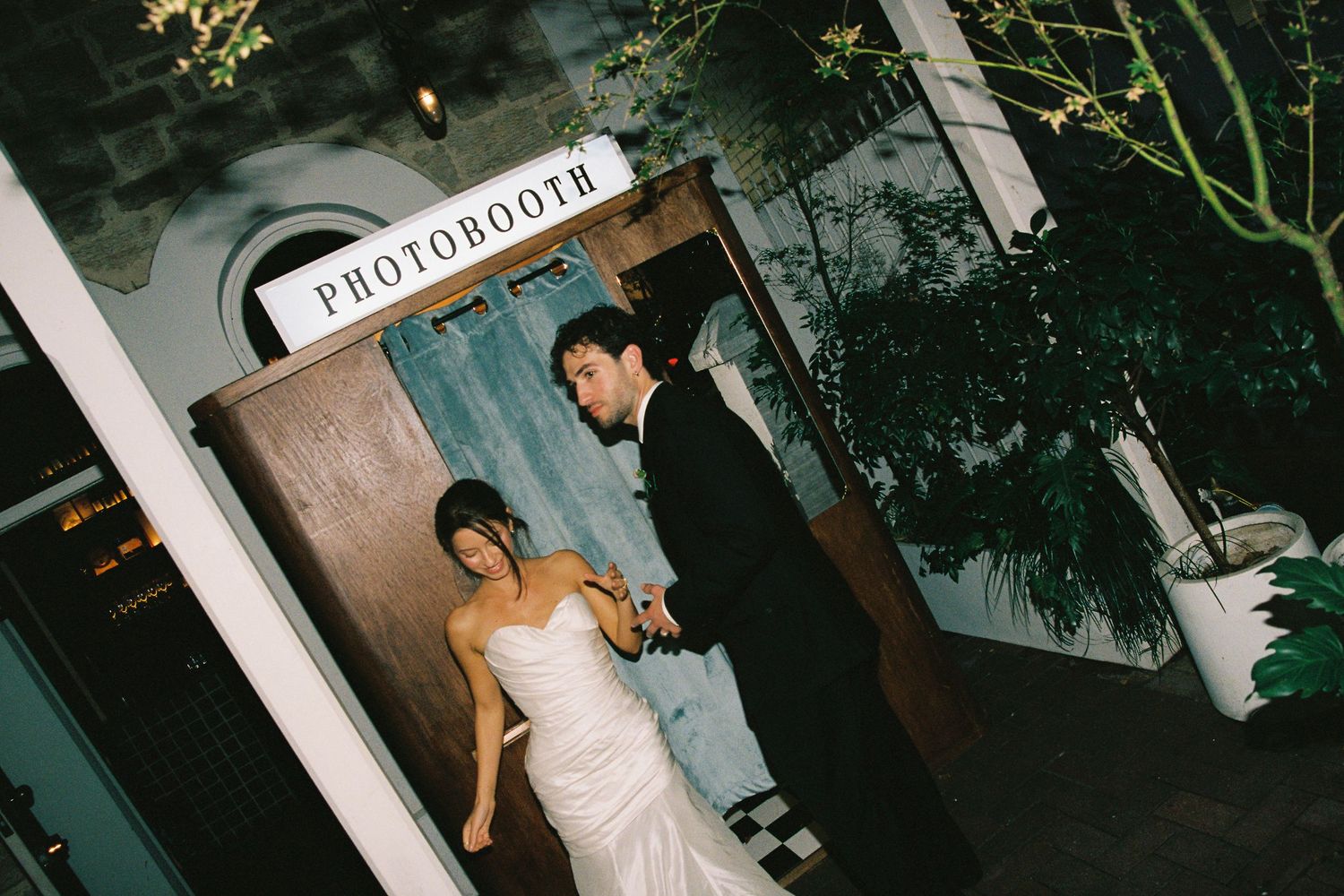 A bride and groom exit a vintage photobooth at their outdoor wedding venue.