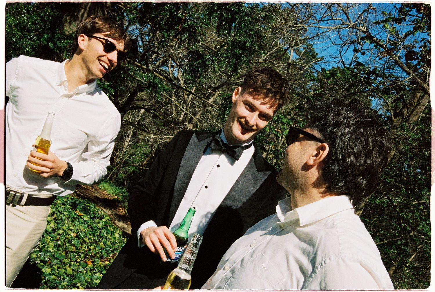 Three wedding guests laughing and drinking champagne outdoors in a garden setting.