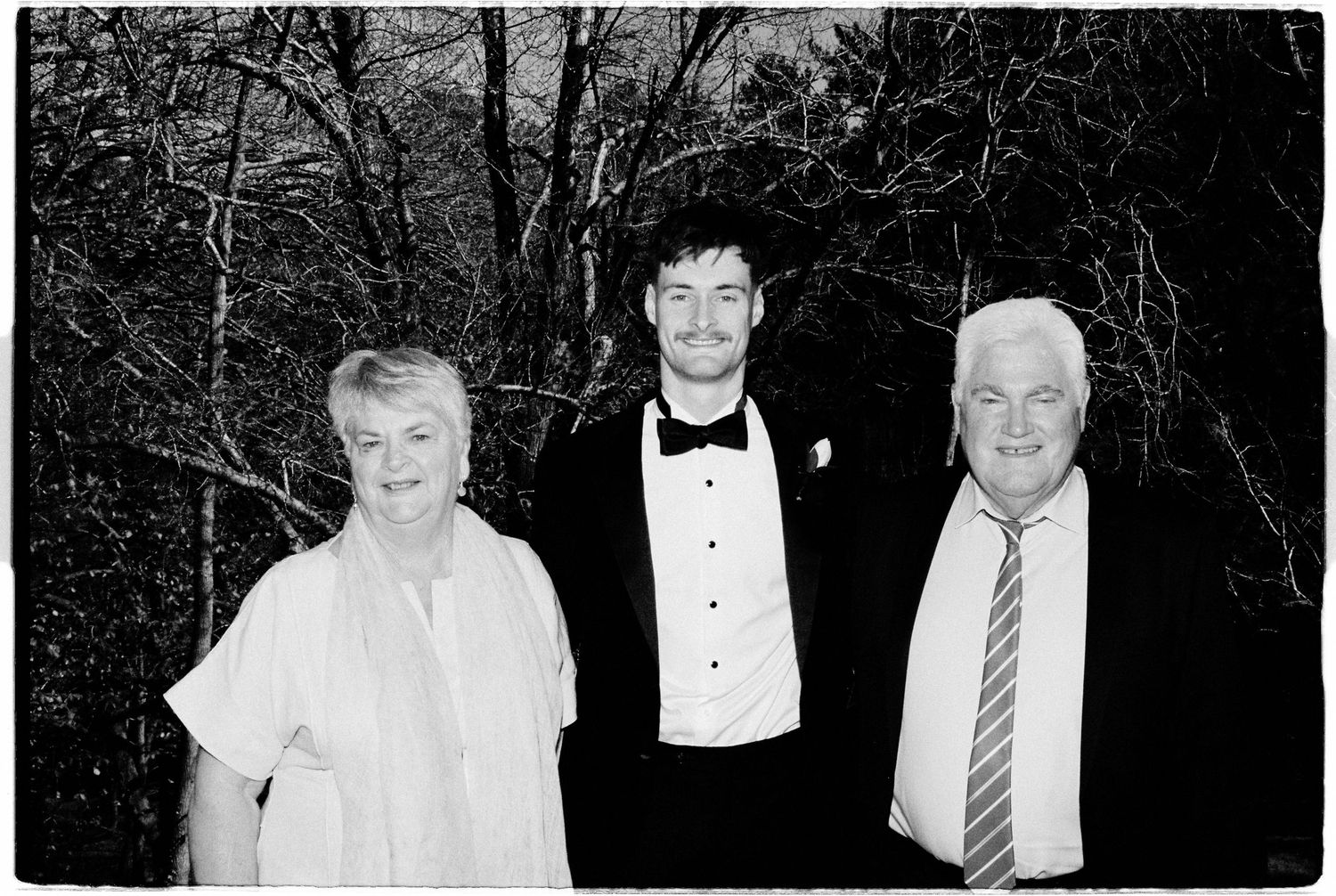Black and white wedding photo of groom in tuxedo posing with two older guests outdoors.