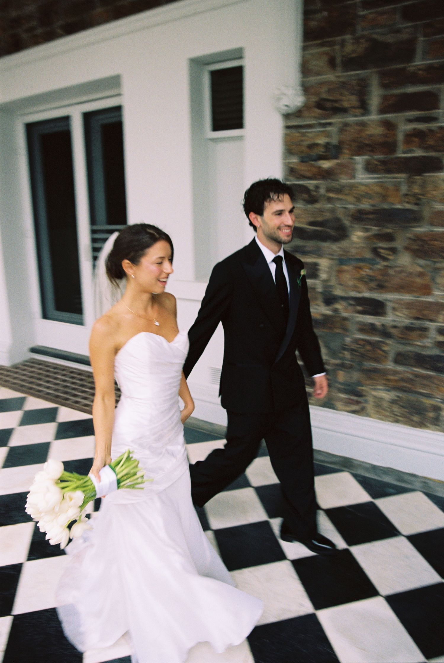 Smiling bride and groom walk together on black and white checkered floor outside stone building.