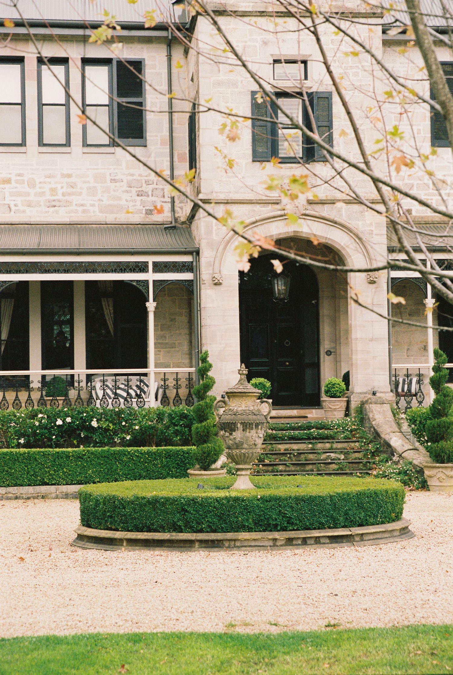 Elegant stone manor house with manicured hedges, ornate urns, and arched black door entrance surrounded by lush gardens.