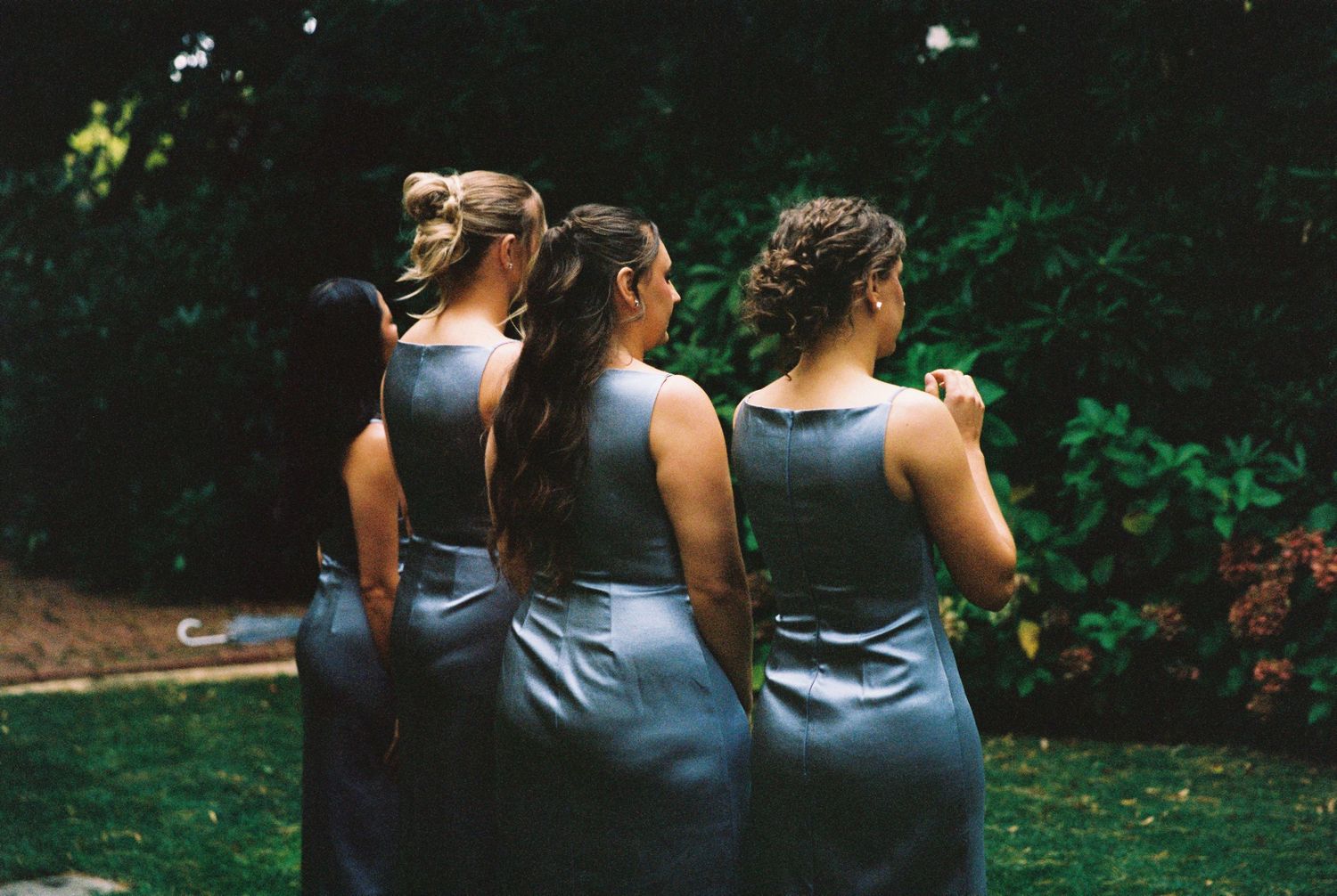 Three bridesmaids in matching slate blue dresses stand together outdoors in a lush garden setting.