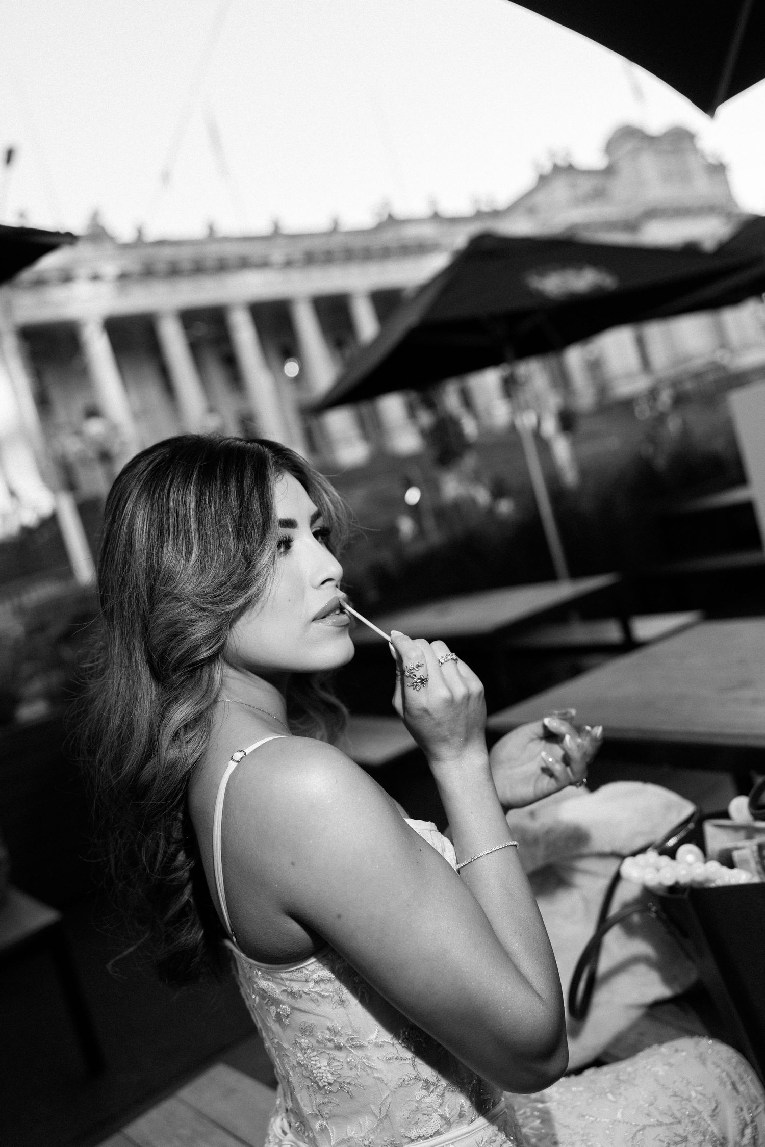 Black and white photo of a bride in lace dress applying lipstick at an outdoor cafe near a historic building.