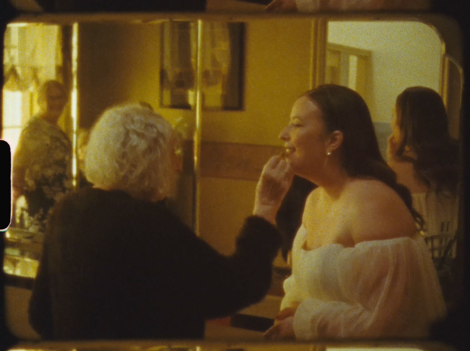 Older woman applying makeup to a young bride in a warmly lit dressing room mirror.