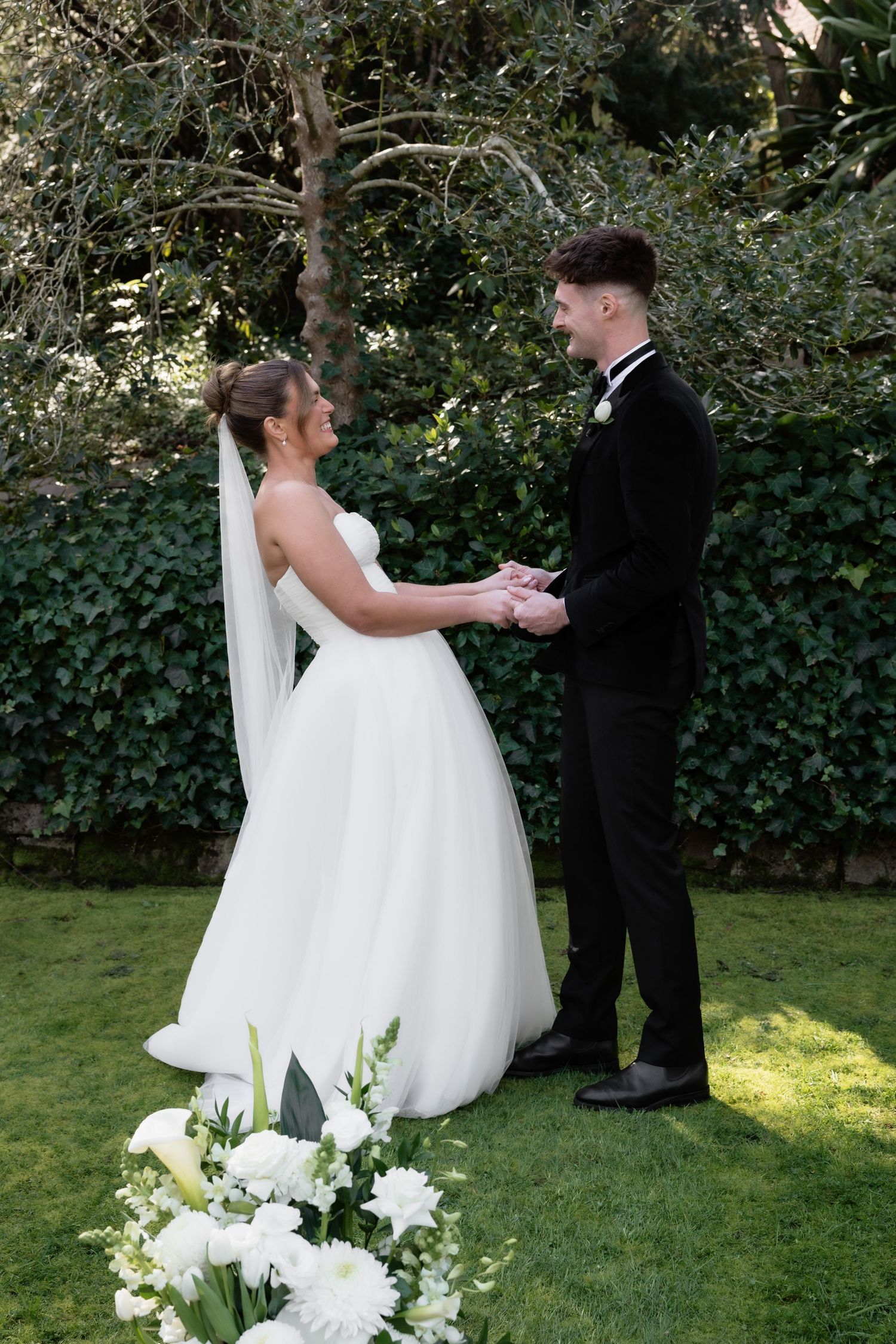 Bride and groom holding hands outdoors on green lawn with white floral arrangement, surrounded by lush garden greenery.