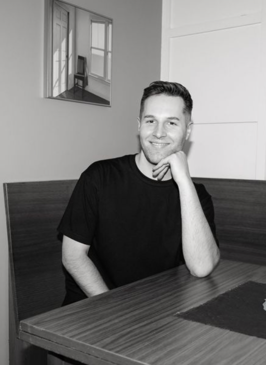 A smiling man in a black t-shirt sits at a wooden table in a modern dining nook, resting his chin on his hand.