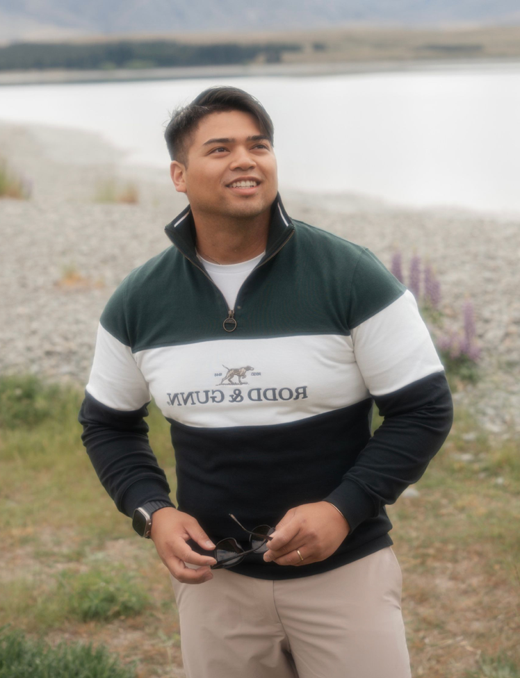 Smiling man wearing a Rodd & Gunn colorblock pullover stands outdoors near a lake with rocky shores.