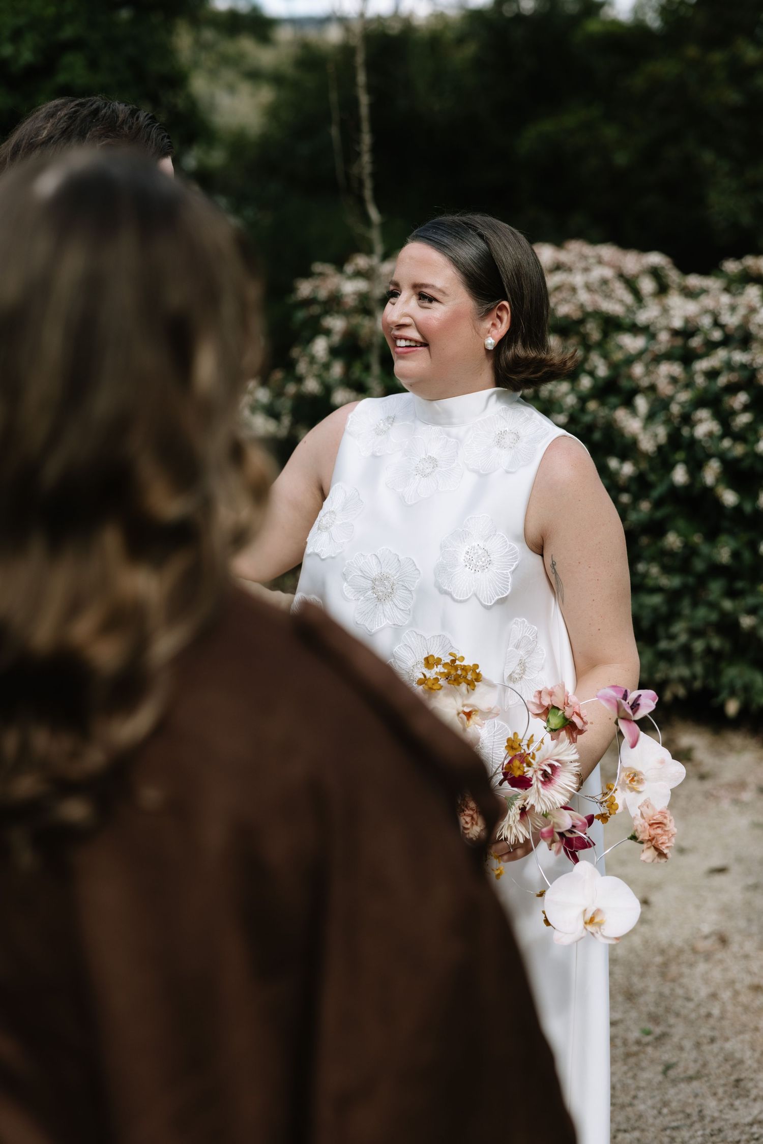 Bride in white floral dress holding colorful bouquet smiles at partner outdoors near blooming garden.