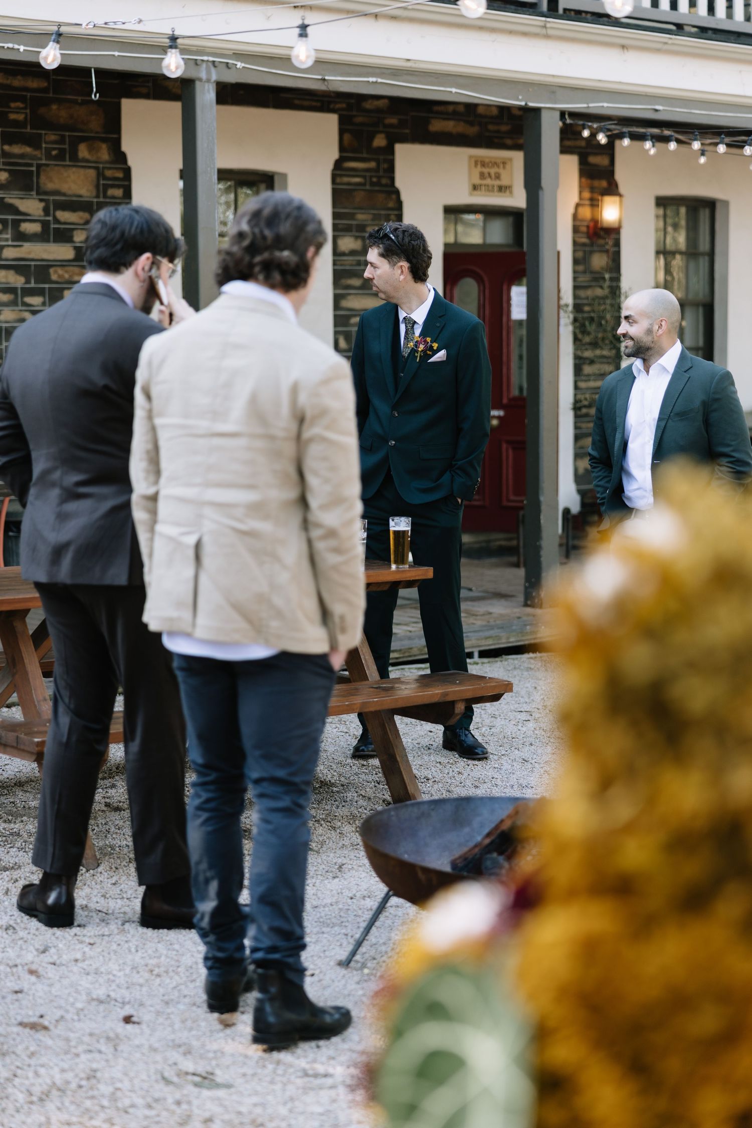 Groom in dark suit with boutonniere socializing with guests outdoors at a rustic wedding venue.