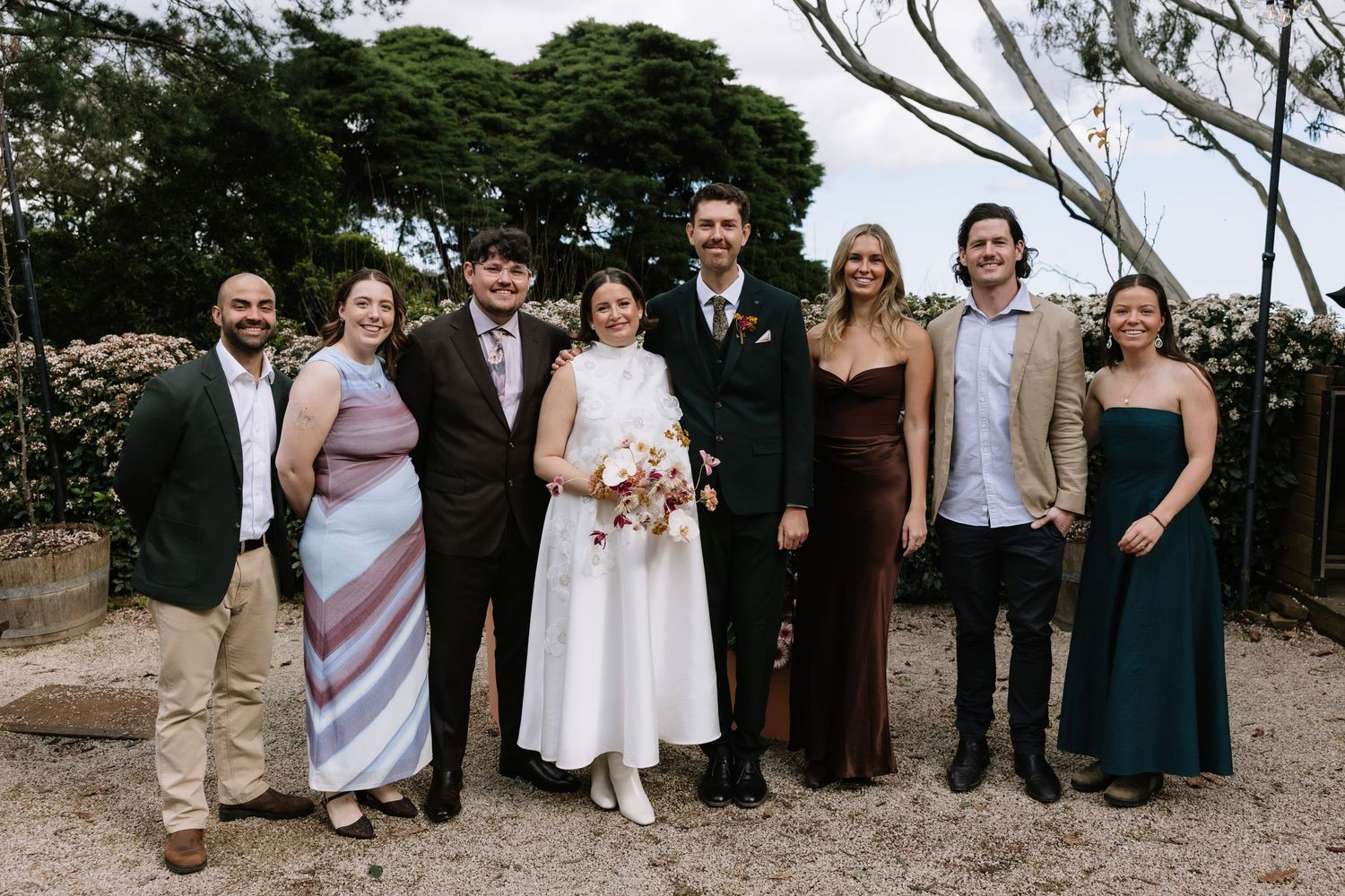 A wedding party of eight people smiling outdoors, with the bride in white and groom in a suit at center.