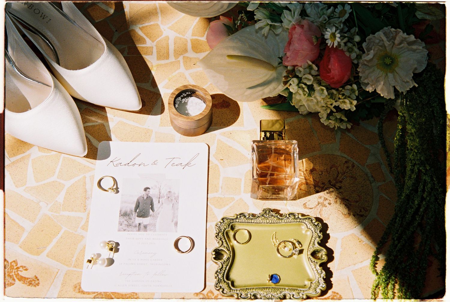 Elegant wedding flat lay featuring bridal shoes, gold rings, perfume bottle, floral bouquet, and invitation on a mosaic surface.