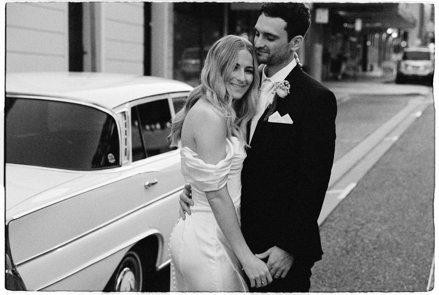 Black and white portrait of smiling newlyweds embracing beside vintage white wedding car on city street.