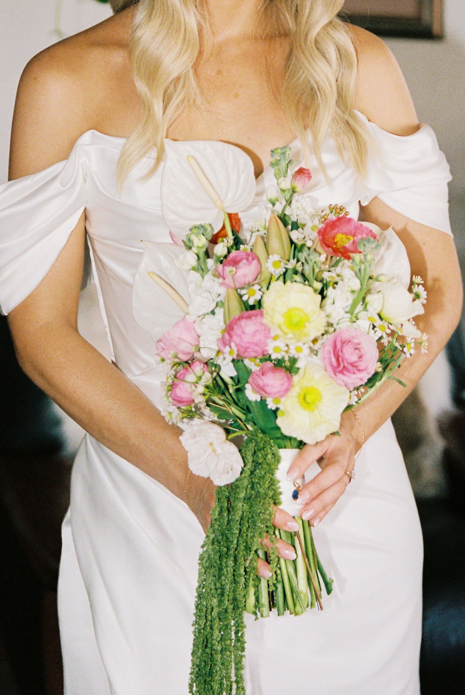 Bride holding a colorful wedding bouquet with pink ranunculus and yellow flowers in a white off-shoulder gown.