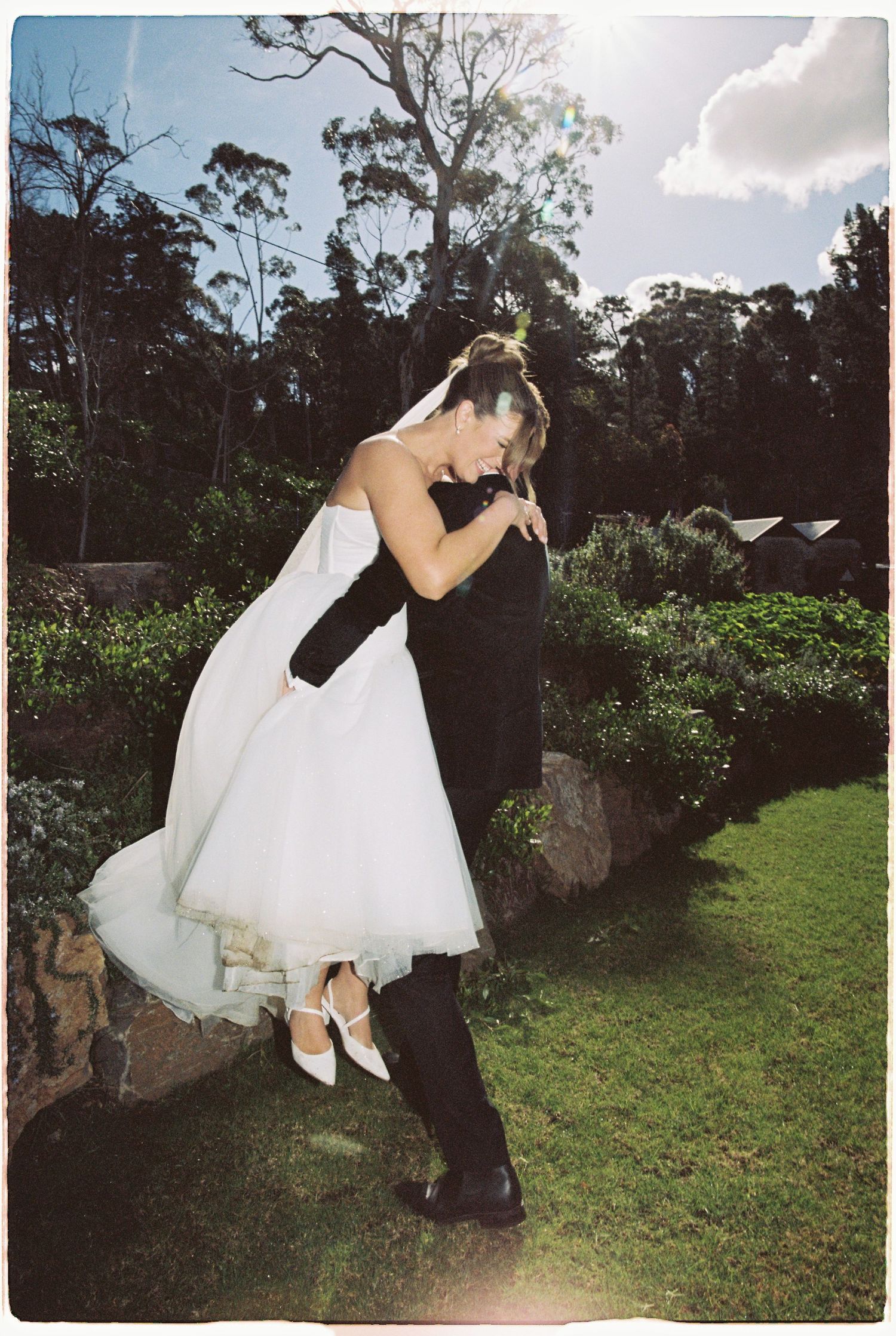Groom lifts bride in white dress outdoors in a sunny garden with trees and blue sky behind them.