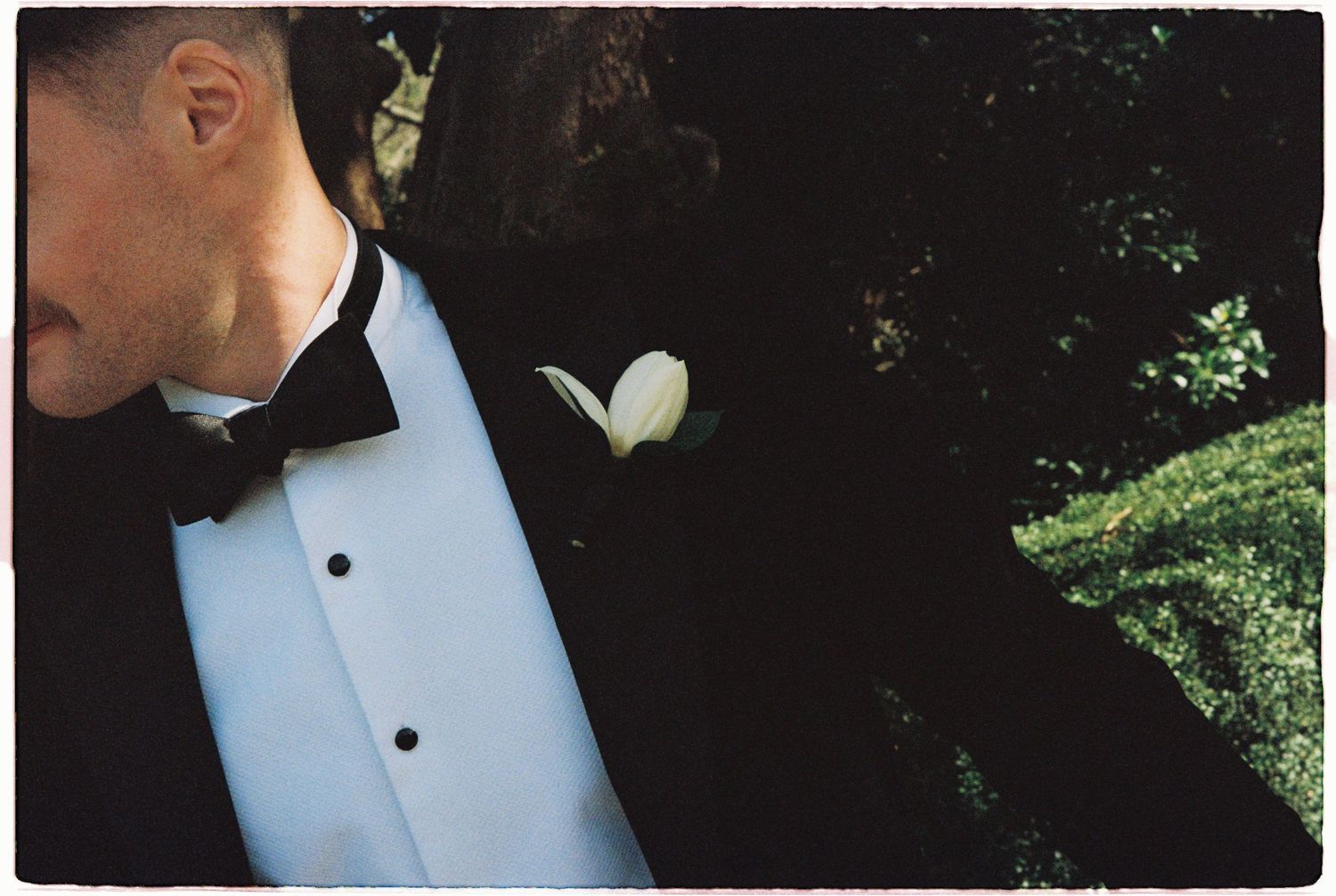 Close-up of groom in black tuxedo with white boutonniere against green garden backdrop.