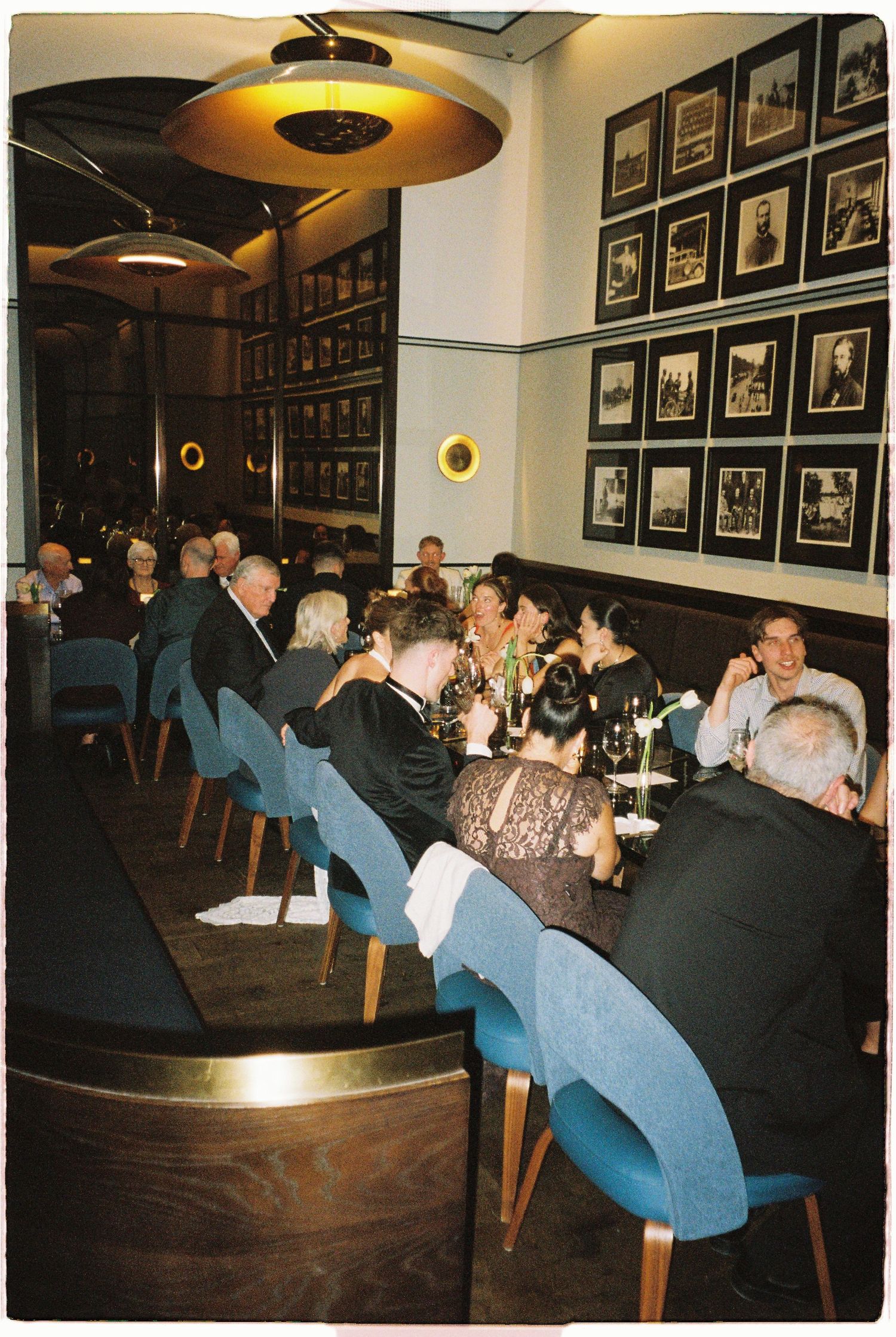 Wedding reception dinner guests seated at long tables in a stylish restaurant with framed photos on walls.