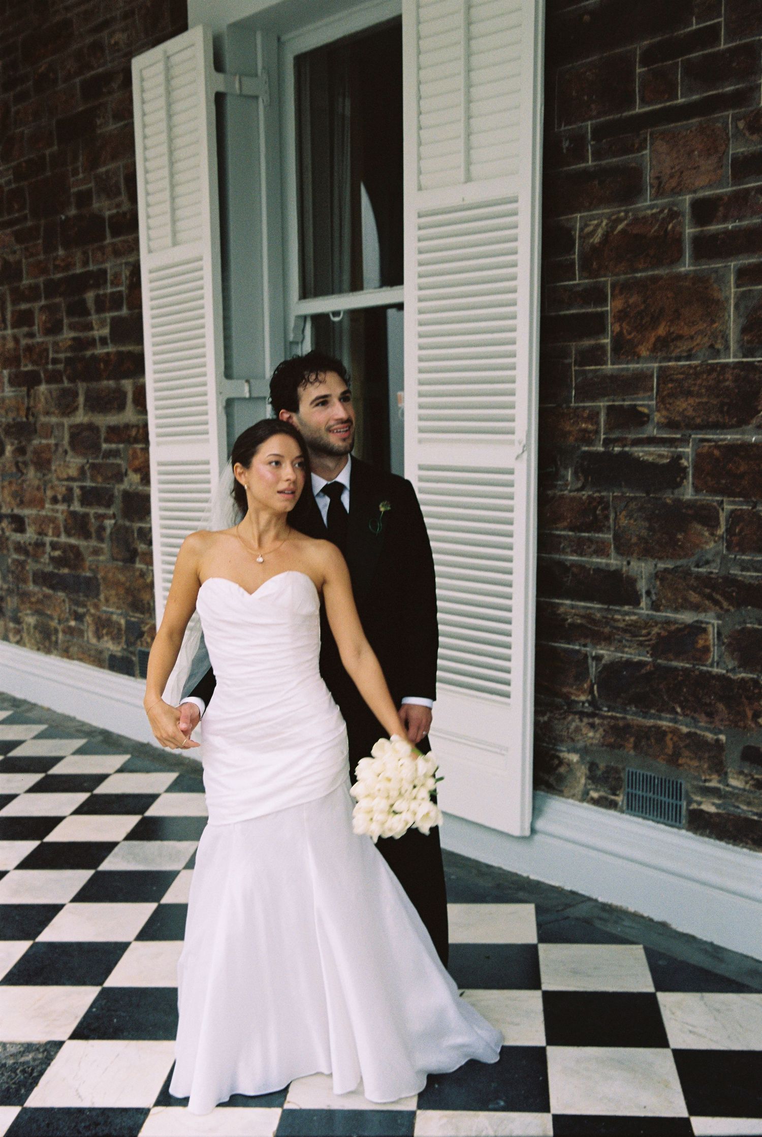 Bride in white strapless gown and groom in black suit pose on checkered floor porch of brick building.