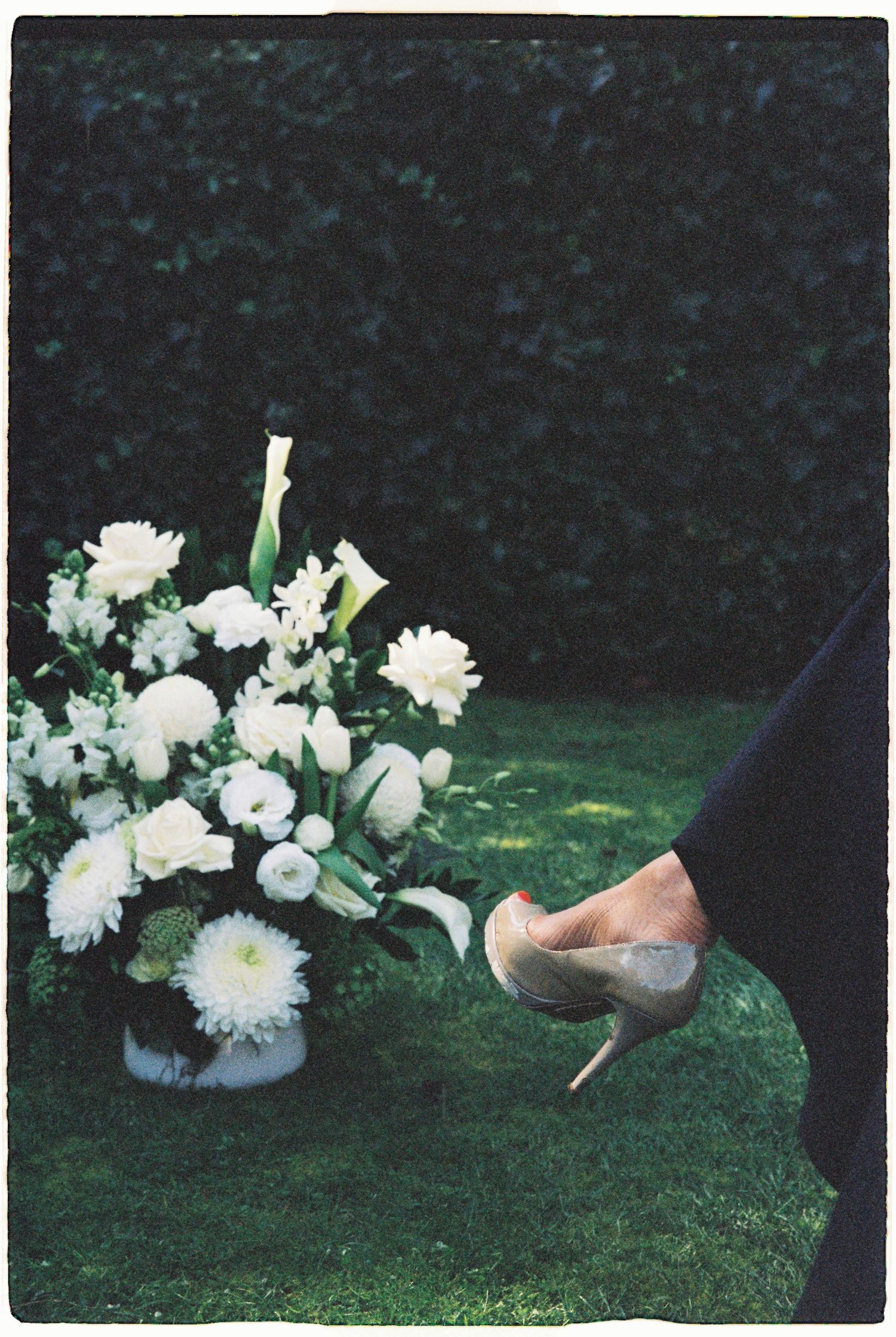 An elegant white floral wedding arrangement on grass beside a woman wearing nude stiletto heels.