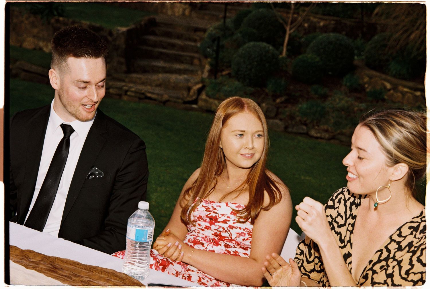 Three guests in formal attire seated at an outdoor wedding reception table conversing together.