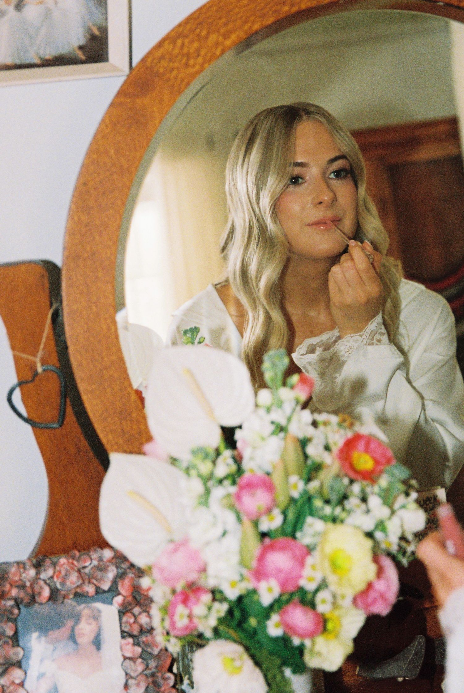 Blonde bride applying lipstick in vintage round wooden mirror surrounded by colorful wedding flowers.