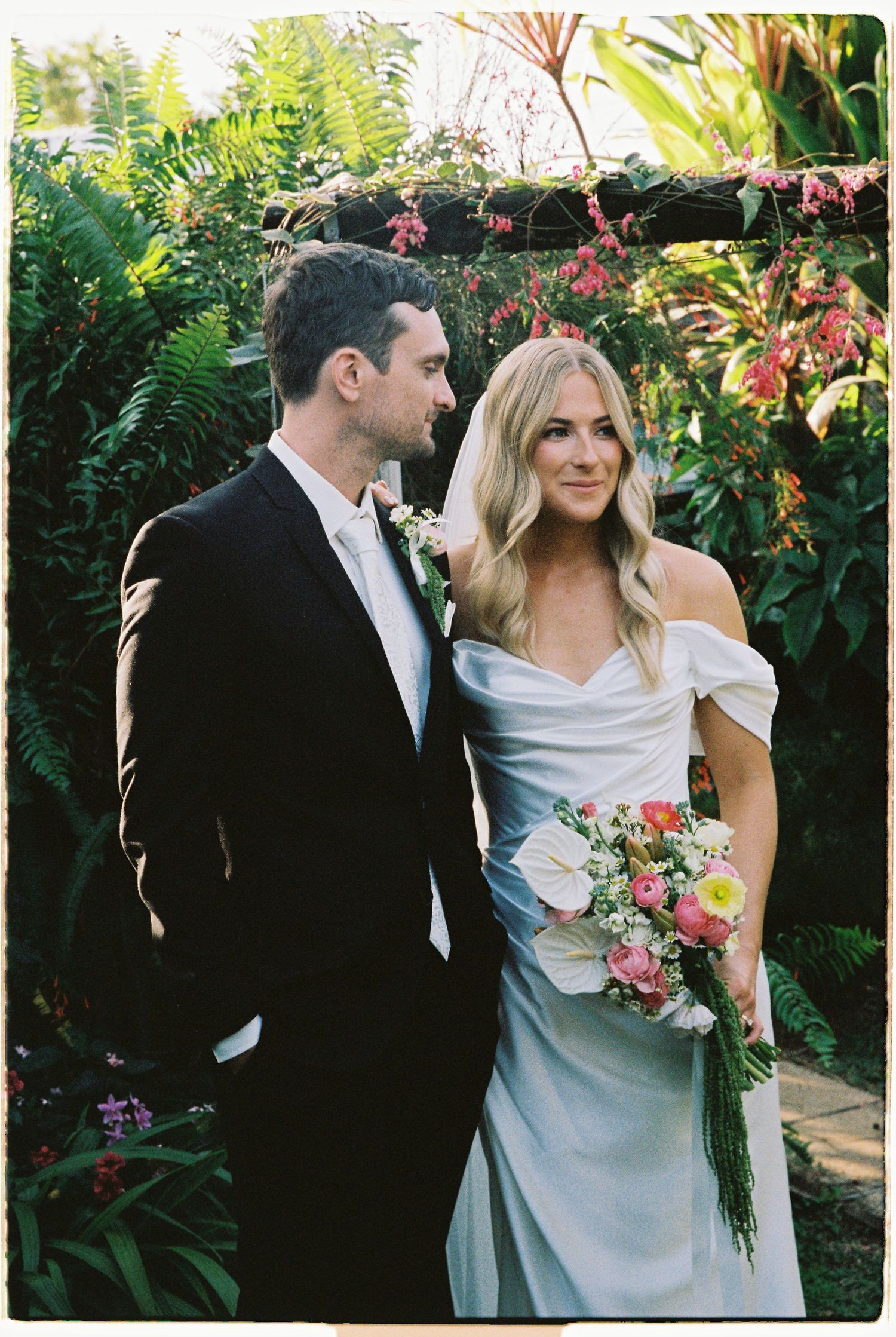 Bride and groom portrait in lush garden, bride holding pink and white bouquet in elegant off-shoulder dress.