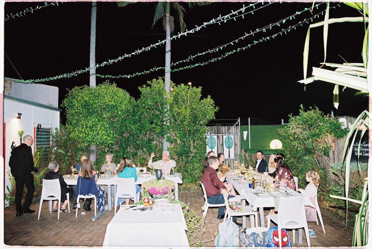 Outdoor evening dinner party with guests dining under string lights surrounded by lush trees at night.