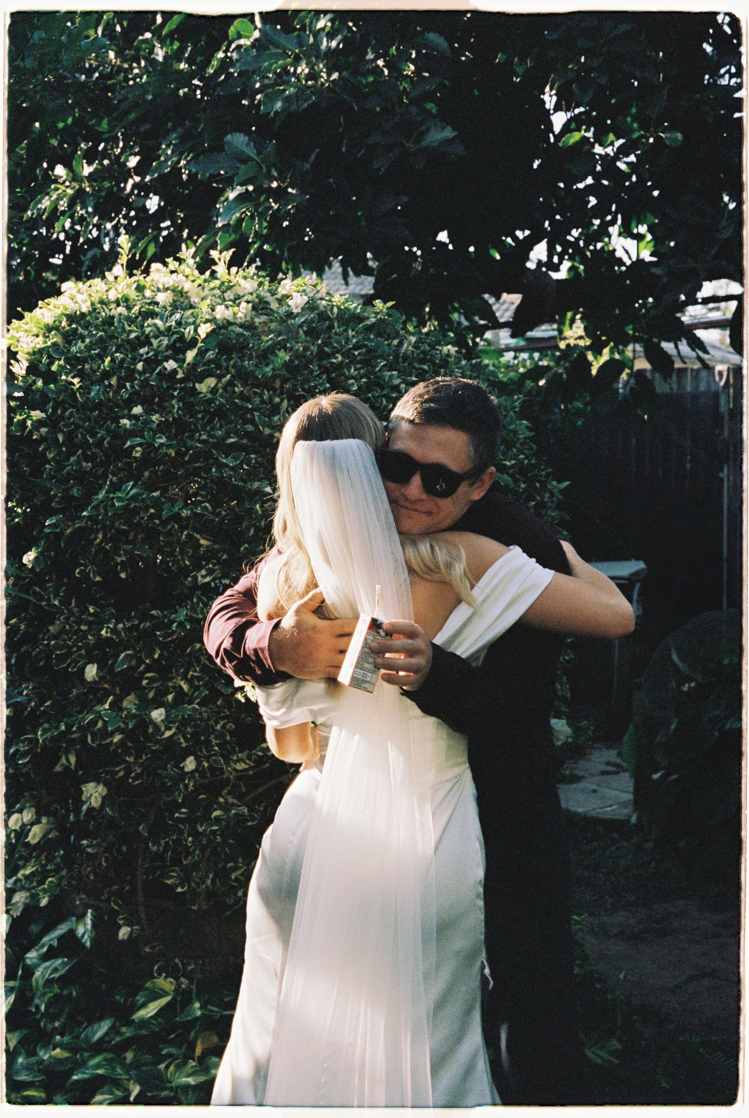 Bride in white gown sharing warm embrace with guest wearing sunglasses in garden setting.